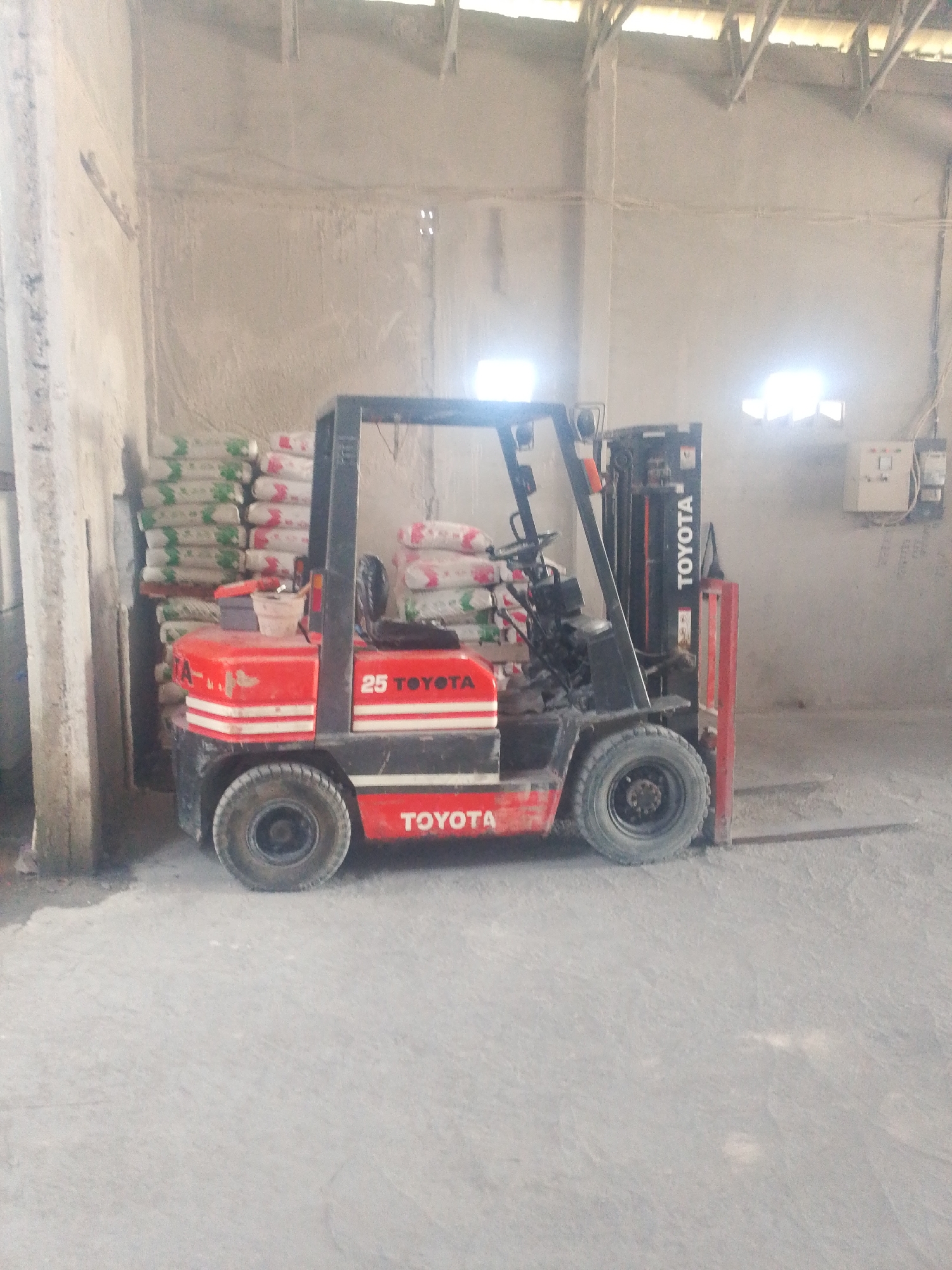 A red and black Toyota forklift is parked indoors on a concrete floor. The forklift is positioned next to stacks of bagged materials, some of which appear to be cement or similar building supplies, and are wrapped in plastic. The forklift has the number "25" visible on its side, along with the "TOYOTA" branding. The interior environment appears to be a warehouse or storage facility, with concrete walls and ceiling. Natural light is entering from openings in the upper part of the structure, creating bright spots and casting shadows. Electrical conduits and a junction box are visible on the far wall. There are no people present in the image.