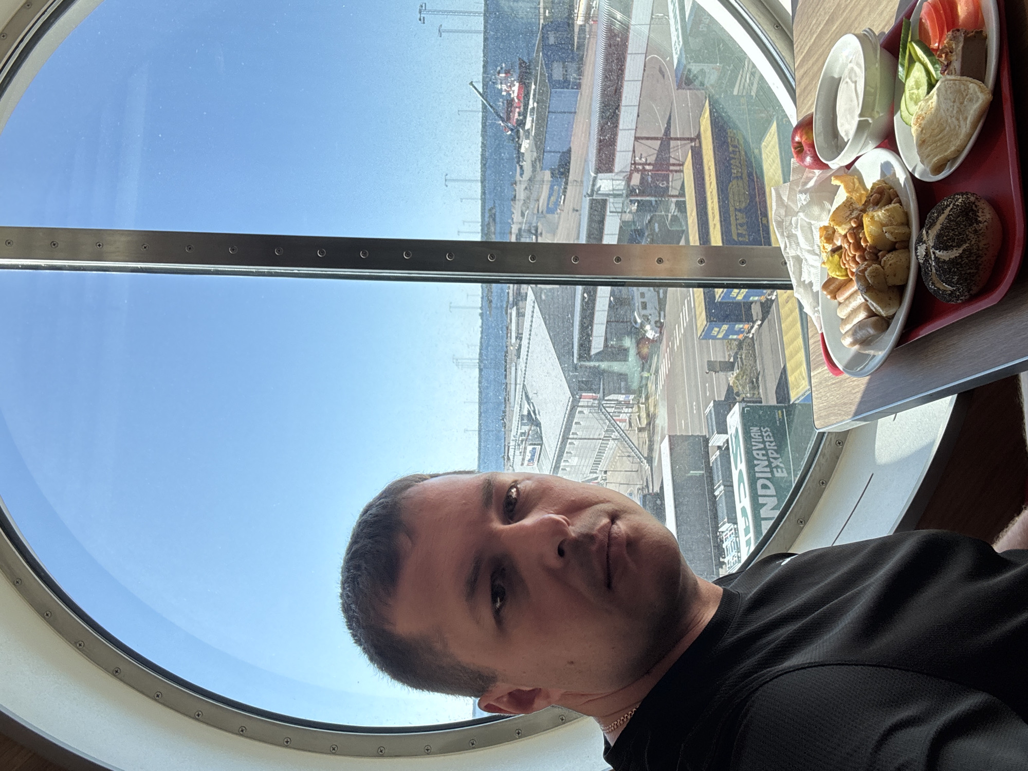 A man sits at a table with a meal in front of him, by a large, circular window. The man is wearing a black t-shirt and a silver chain. He is looking towards the camera. On the table, a red tray holds a plate with sausages, beans, and potatoes, a bread roll, a small plate with a bread roll, cucumber slices, tomato slices, and a piece of meat. There is also a red apple and a bowl of white yogurt. Through the window, a port is visible with industrial buildings, shipping containers, and a red and white ship in the water. The sky is clear and blue. This scene is likely in the city of Karlskrona, Sweden, as suggested by the "Scandinavian Express" shipping container, a known ferry operator.