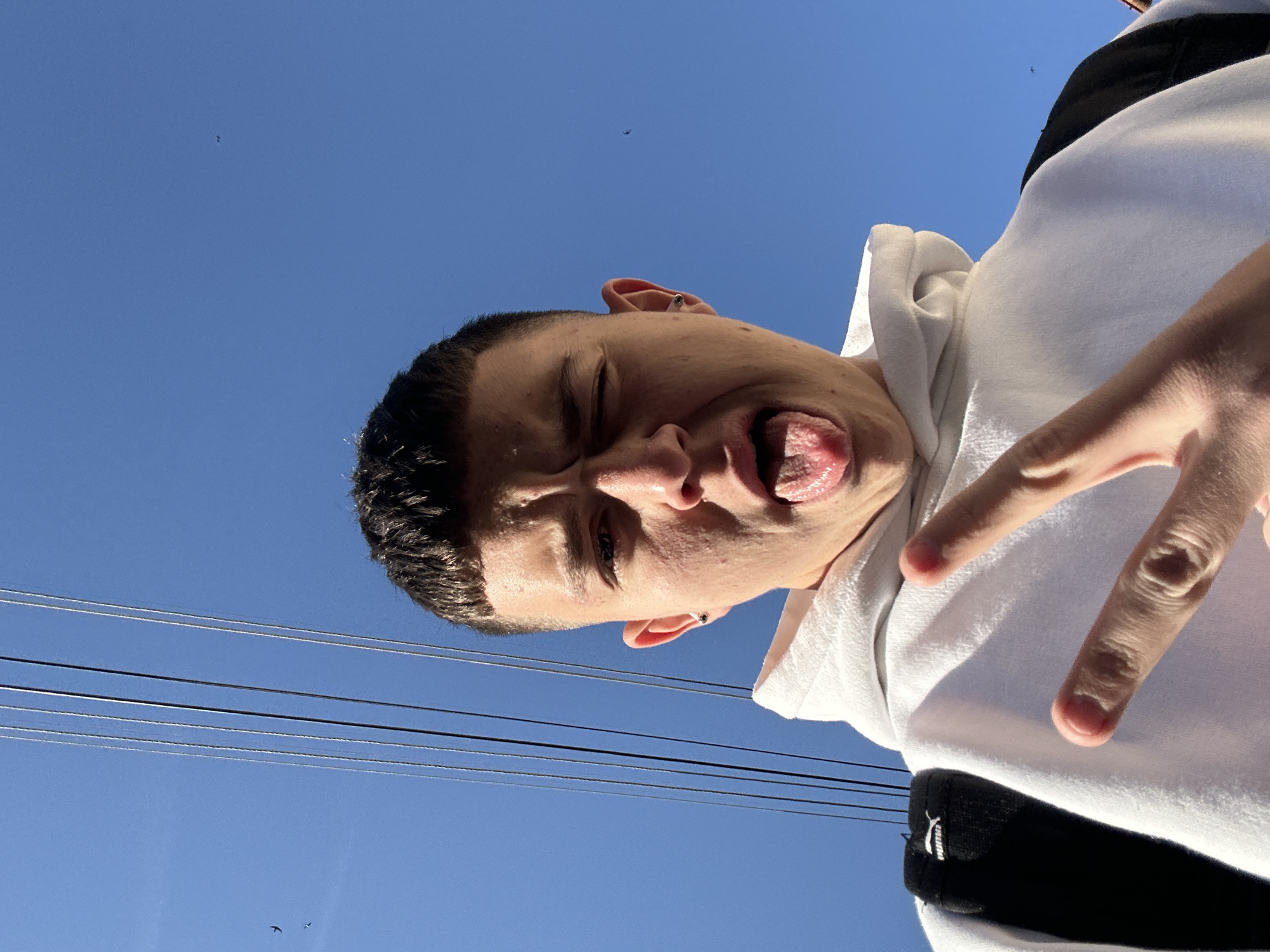 A low-angle, upward-tilted selfie of a young man with short, dark hair. His eyes are closed, his tongue is protruding, and he is making a peace sign with his right hand. He is wearing a white hoodie with a black band. Above him is a clear blue sky with electrical wires visible. Two small birds are discernible in the upper left corner of the frame. The specific location in Zlatograd, Bulgaria, is indeterminable from the given visual information.