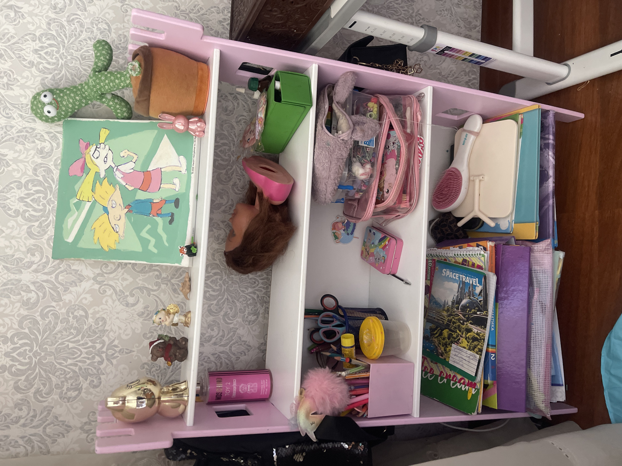 The image depicts a pink, multi-tiered shelf unit filled with various items. The top shelf holds a gold-colored figurine, a small teddy bear, a figurine of an angel with golden wings, a small brown object, a framed picture featuring cartoon characters Arnold and Helga Pataki from the show Hey Arnold!, and a plush green cactus in a terracotta pot with a small white flower. To the left of the shelf unit, a black sequined garment is visible.
The second shelf contains a pink bottle labeled "Moschino Toy 2 Bubble Gum," a mannequin's head with brown hair and a pink face, a green rectangular box, and a small bottle with a green cap.
The third shelf is organized with a pink container holding several pens and pencils, a fluffy pink pom-pom accessory, a yellow-capped container, and a blue metal container filled with scissors and more pens. To the right of these items are two small pink containers, one of which is open and contains various crafting supplies, and a pink tin box with a pen lying next to it.
The fourth shelf contains several stacks of notebooks and books. One notebook is green and features a "Space Travel" title. Below it is a purple binder.
The bottom shelf holds a stack of notebooks and folders, a white hairbrush with a pink handle, and a light-colored cosmetic palette. To the right of the shelf unit is a white adjustable desk leg with colored markers. The background consists of a grey patterned wallpaper. The floor is made of polished wood.