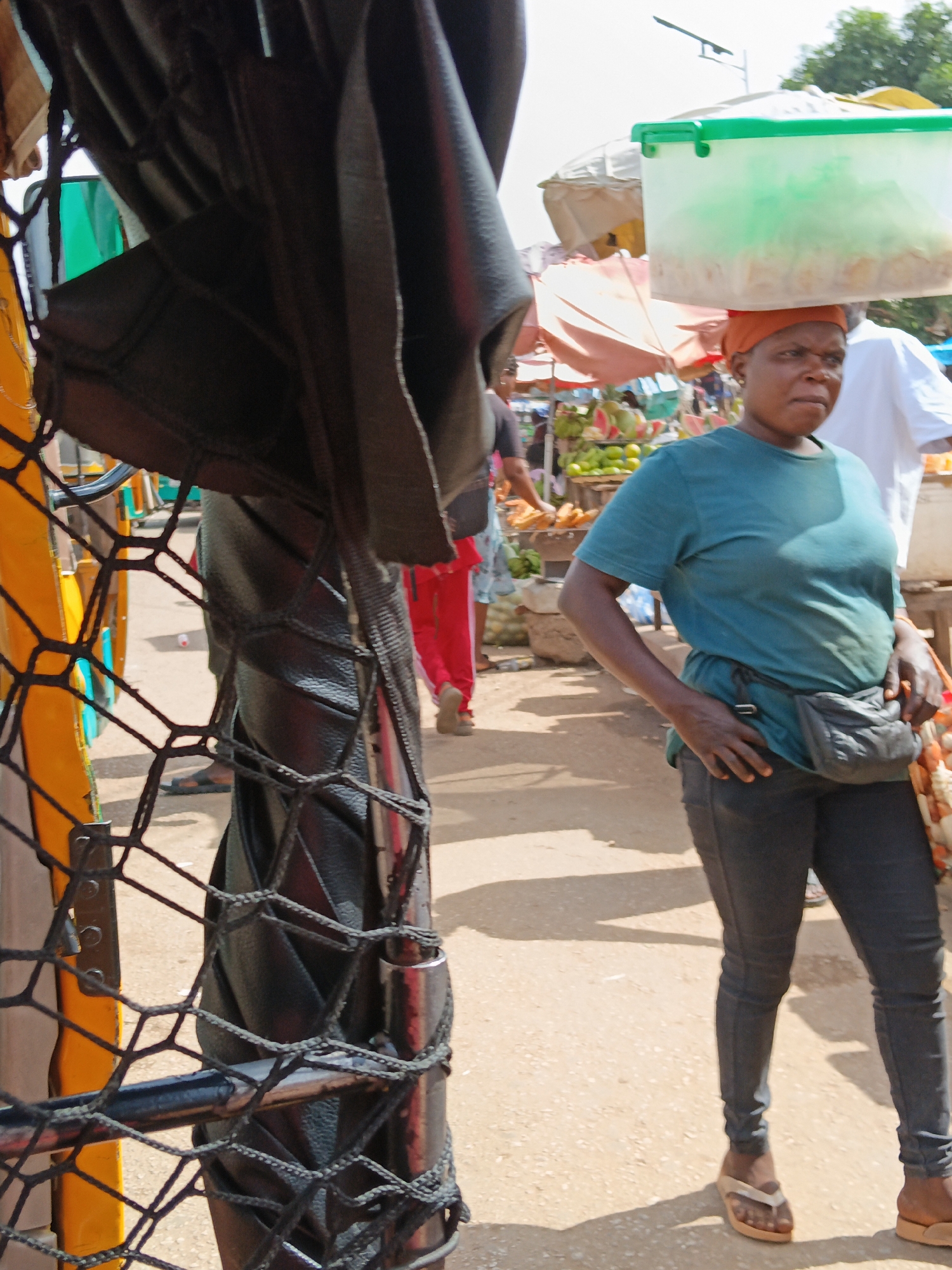 A woman wearing a teal t-shirt and black pants carries a light green plastic container on her head while walking through an outdoor market. She has an orange headscarf and a black fanny pack around her waist. In the background, other market-goers and stalls with fruits and vegetables are visible. The ground is dusty and unpaved, suggesting an urban environment in a city like Unknown, Unknown. The foreground shows a partial view of a yellow and black structure, possibly a vehicle or market stall, with black netting.