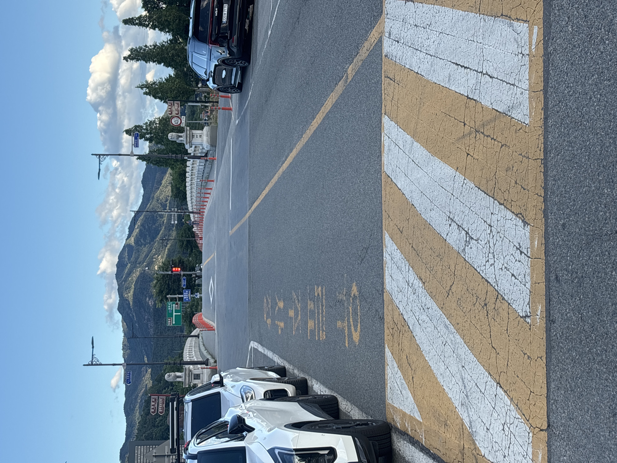 The image captures a street scene in Gyeongju-si, South Korea, on a clear day with a blue sky dotted with white clouds. In the foreground, a crosswalk with yellow and white stripes is visible. The road is paved and has a yellow dividing line. Several cars are parked on the side of the road; two white SUVs are on the left, and a black Kia Carnival is on the right. Orange traffic cones line the edge of the road. In the background, a bridge with white railings and red accents spans across, leading towards a mountainous landscape. Streetlights and road signs are present along the road and bridge. One road sign, in green, indicates directions to "Bomun IC" and "Gyeongju." Another sign, with Korean characters, is painted in yellow on the asphalt, partially obscured by the crosswalk. The overall environment suggests a well-maintained urban area with natural scenery.