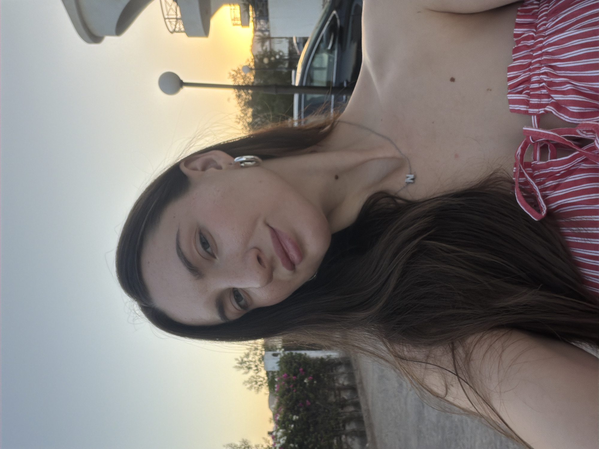 A young woman with long, dark brown hair, wearing a red and white striped off-the-shoulder top and a silver necklace with a pendant, poses for a selfie outdoors at sunset in Sharm El Sheikh, Egypt. The background features a paved walkway, a hedge with pink and white flowers, a street lamp, a dark-colored car, and a building. The sky is a gradient of pale blue and orange, indicative of the setting sun.