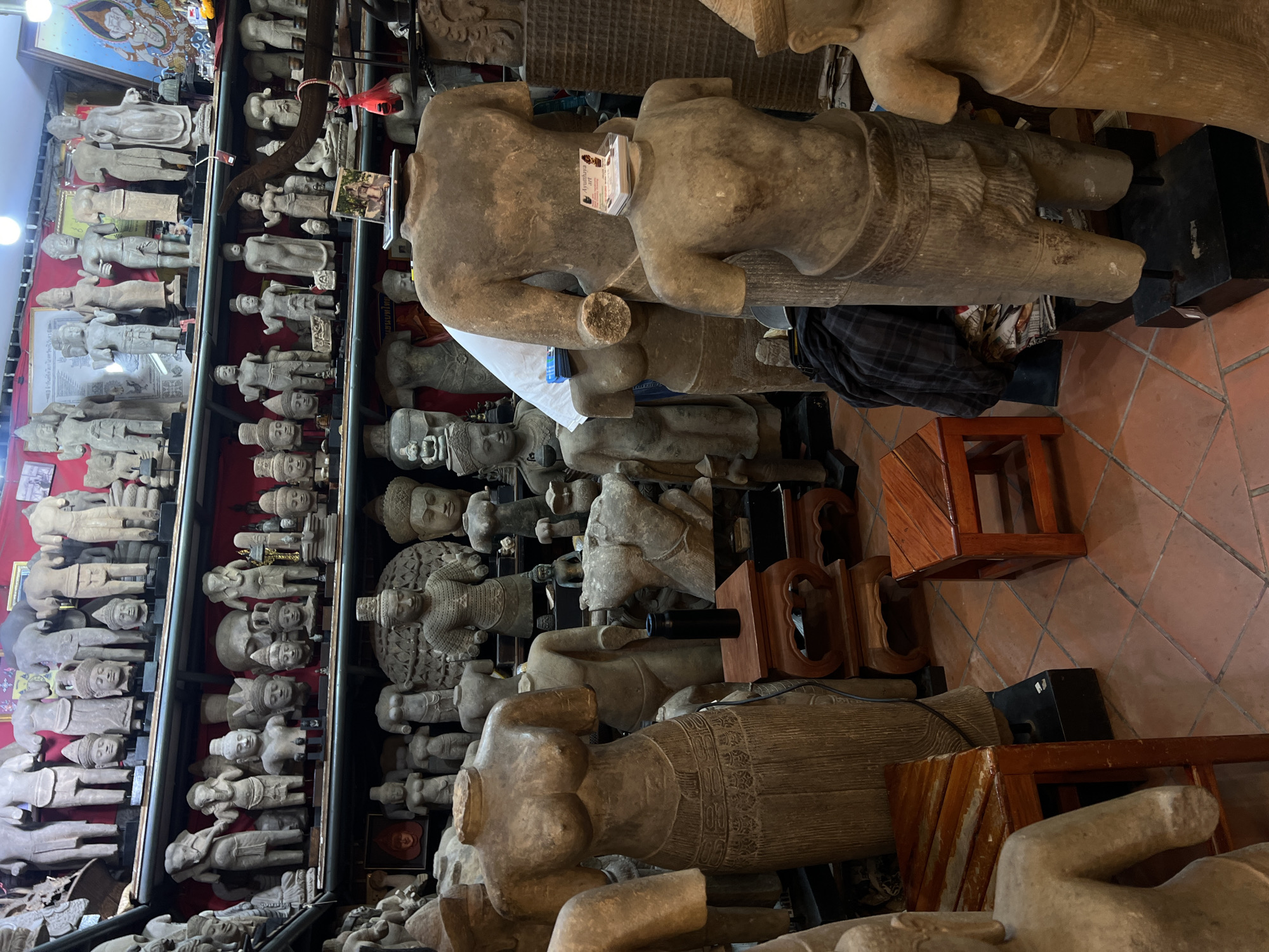 The image shows a densely packed room filled with stone statues, primarily torsos and heads, displayed on shelves and the floor. The scene appears to be a storage area or shop selling these artifacts. The majority of the statues are gray or beige in color, suggesting they are made of stone or a similar material. Some have been damaged or are incomplete. The location is likely in Bangkok, Thailand, due to the presence of numerous statues with traditional Thai cultural and religious motifs. There are also wooden stands and stools scattered around the room. The floor is tiled with reddish-brown tiles. The lighting in the room is somewhat dim, with a mix of natural and artificial light sources.