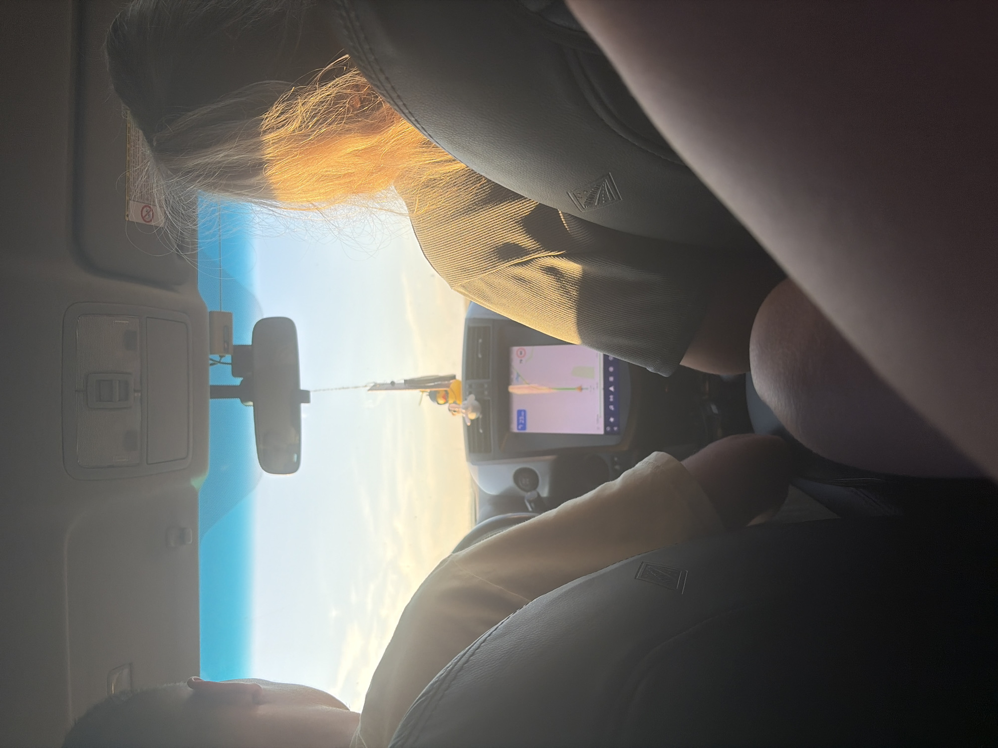 The image shows the interior of a car, viewed from the back seat, with two passengers visible. The person in the front passenger seat, a woman with long blonde hair, is illuminated by golden hour sunlight. To her left, partially visible, is another passenger. The car's dashboard features a navigation screen displaying a route. Outside the car, the sky is clear and bright, suggesting a daytime drive. The location is identified as Belogorki, Russia.