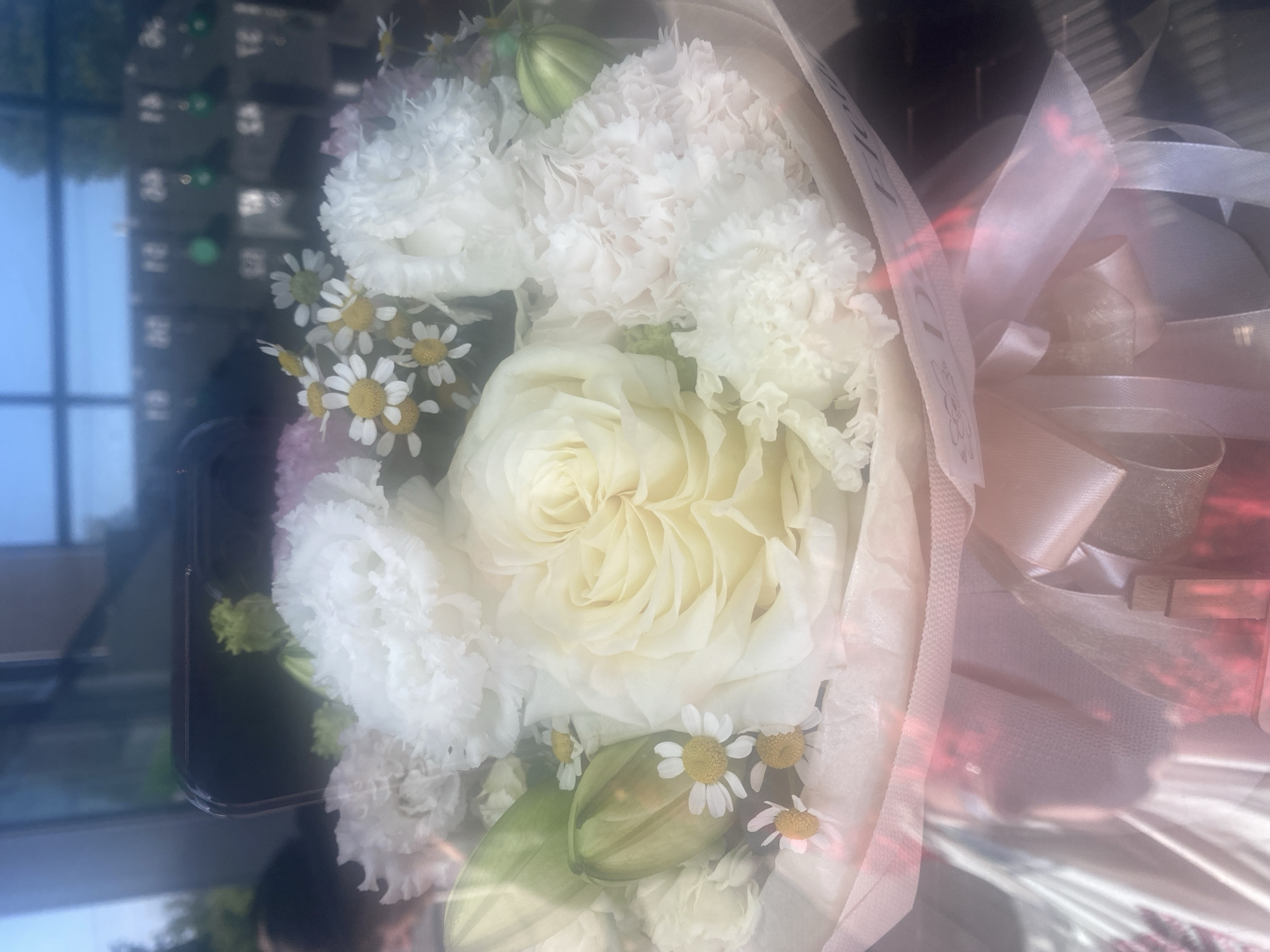 A bouquet of white flowers is displayed, featuring a large, pale yellow rose as the central focus. Surrounding it are various white blooms with ruffled petals, possibly carnations or lisianthus, interspersed with small, daisy-like flowers. A green bud, possibly from a lily, is also visible. The bouquet is wrapped in sheer, pale pink and white material, tied with satin ribbons. The overall impression is one of delicate beauty and freshness.
The image appears to be taken indoors, possibly in a studio or a room with large windows, given the reflections seen in the background. The lighting suggests it could be daytime, with natural light illuminating the bouquet. There are reflections of what appear to be buildings and a bright sky in the background, indicating an outdoor environment is visible through a window. The mood conveyed is serene and celebratory.
The scene is centered on the floral arrangement, with no people prominently visible. The flowers themselves are the primary subjects. The presence of ribbons and wrapping suggests the bouquet is prepared for gifting or an event.
There is some faint, partially obscured text visible on one of the ribbons, with what looks like "LUDOVIK" printed on it, along with some decorative elements. The text is not entirely clear due to the resolution and the angle of the ribbon.
The location context provided is Novi Petrivtsi, Ukraine, though there are no direct visual cues in the image to confirm this specific location. The general setting appears to be a well-lit interior with a view to the outside.