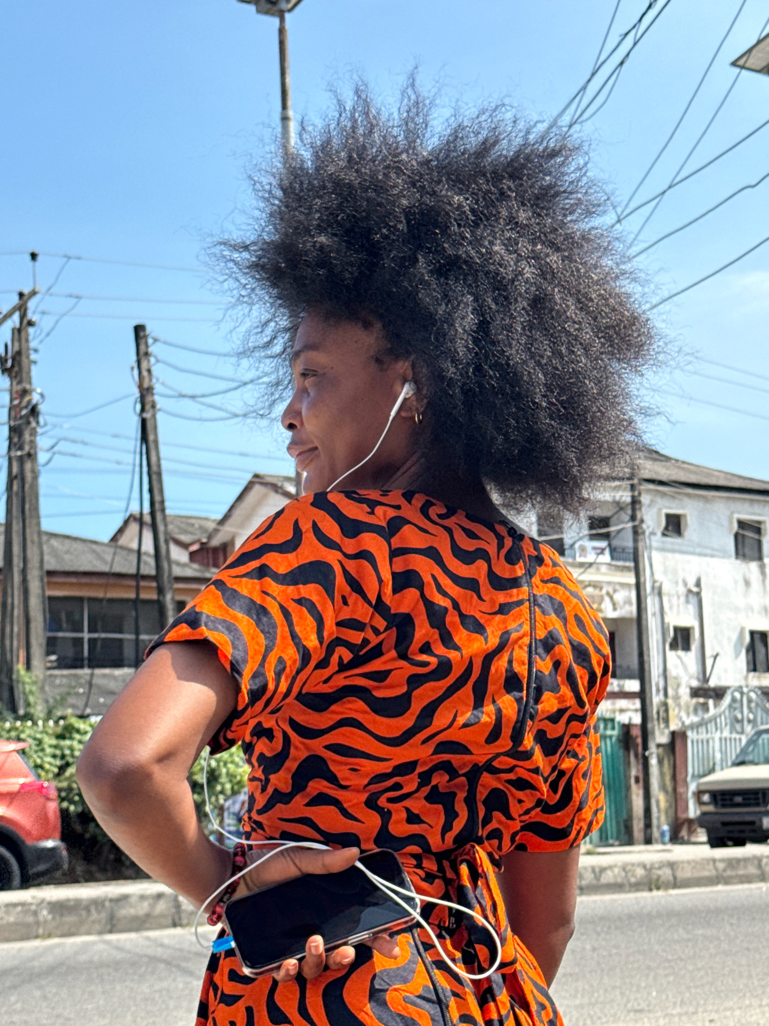 A woman with voluminous, dark, curly hair is shown from the back, looking to her left in an urban outdoor setting. She is wearing an orange and black tiger-striped short-sleeved dress and white wired earbuds. In her right hand, she holds a smartphone, with the wire from the earbuds looped around her fingers and the phone. Her left arm is bent at the elbow, with her hand on her hip. The background includes a bright blue sky, utility poles with numerous wires, and buildings with white and grey facades, one with visible windows. A red vehicle is partially visible on the left, and a light-colored vehicle is parked on the right. The scene appears to be a street in Ado, Nigeria.