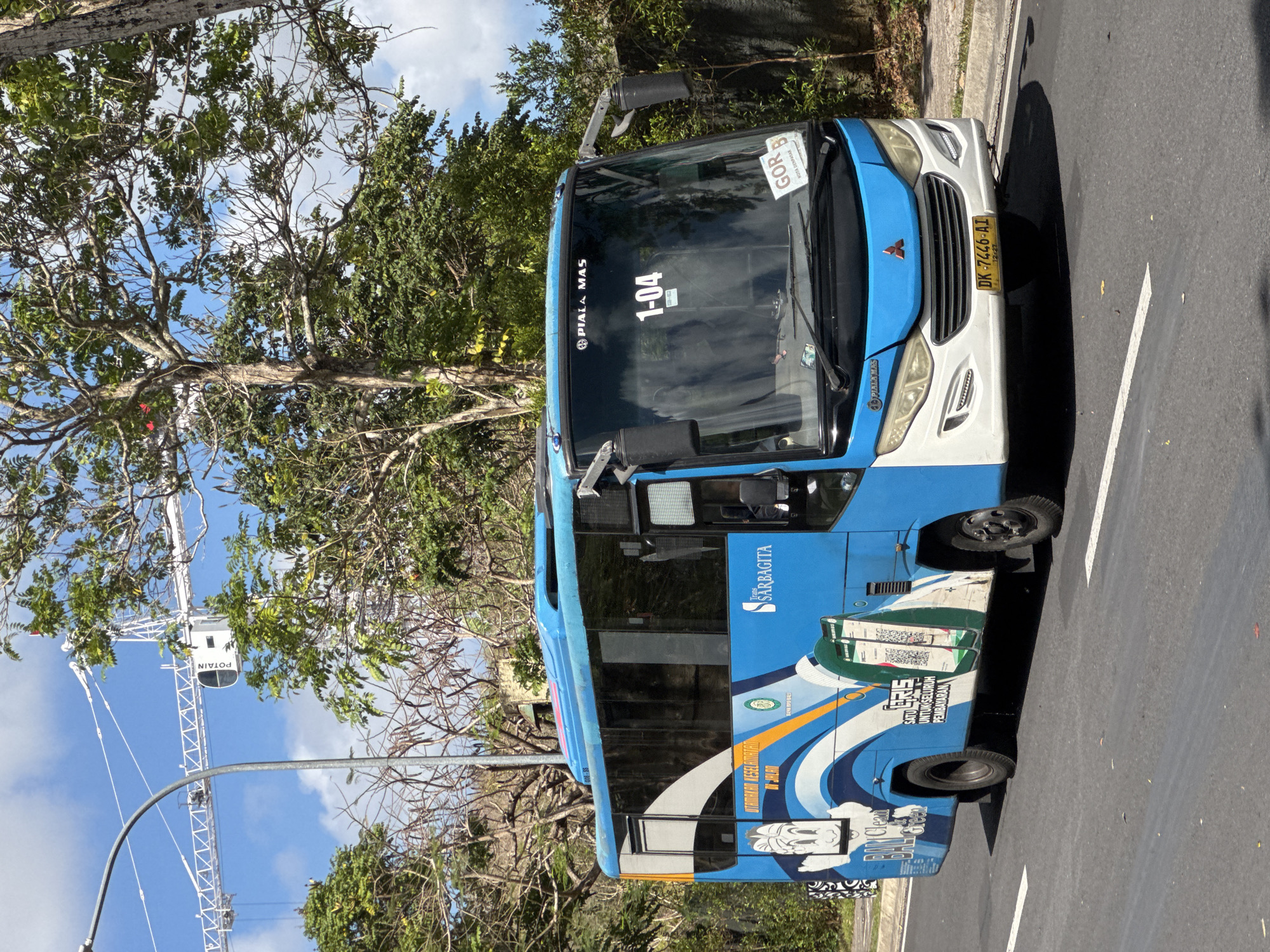 A blue and white bus with the name "Trans Sarbagita" is visible from the side and front. The bus has "1-04" displayed on the windshield. The license plate reads "DK 7446 AI". The bus is on a paved road with white lane markings. In the background, there are trees, a construction crane, and a stone wall. The sky is blue with some clouds. The bus appears to be in motion. The location is Kuta Selatan, Indonesia.