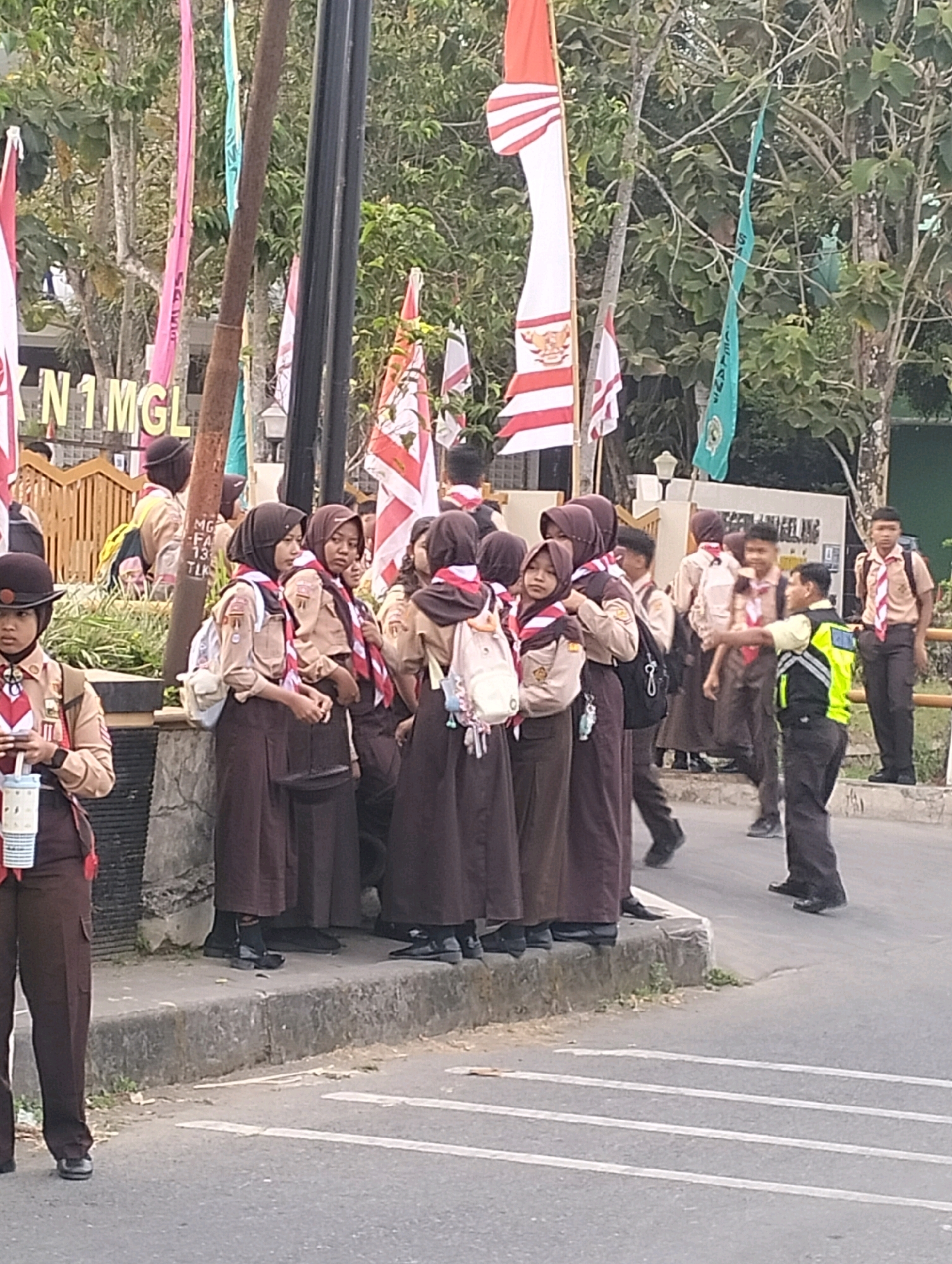 The image shows a group of uniformed students, likely scouts, standing on a sidewalk. They are wearing brown uniforms, scarves, and some have backpacks. The background includes trees, flags, and a building with signage that reads "N1MGL" and partially visible text in Indonesian. A man in a high-visibility vest is also present, interacting with the students. The scene appears to be outdoors, on a street with white lines painted on the asphalt, suggesting it might be a road or a designated area. The presence of the "N1MGL" signage and the Indonesian text strongly suggests the location is in the city of Magelang, Indonesia, as "MGL" is a common abbreviation for Magelang.