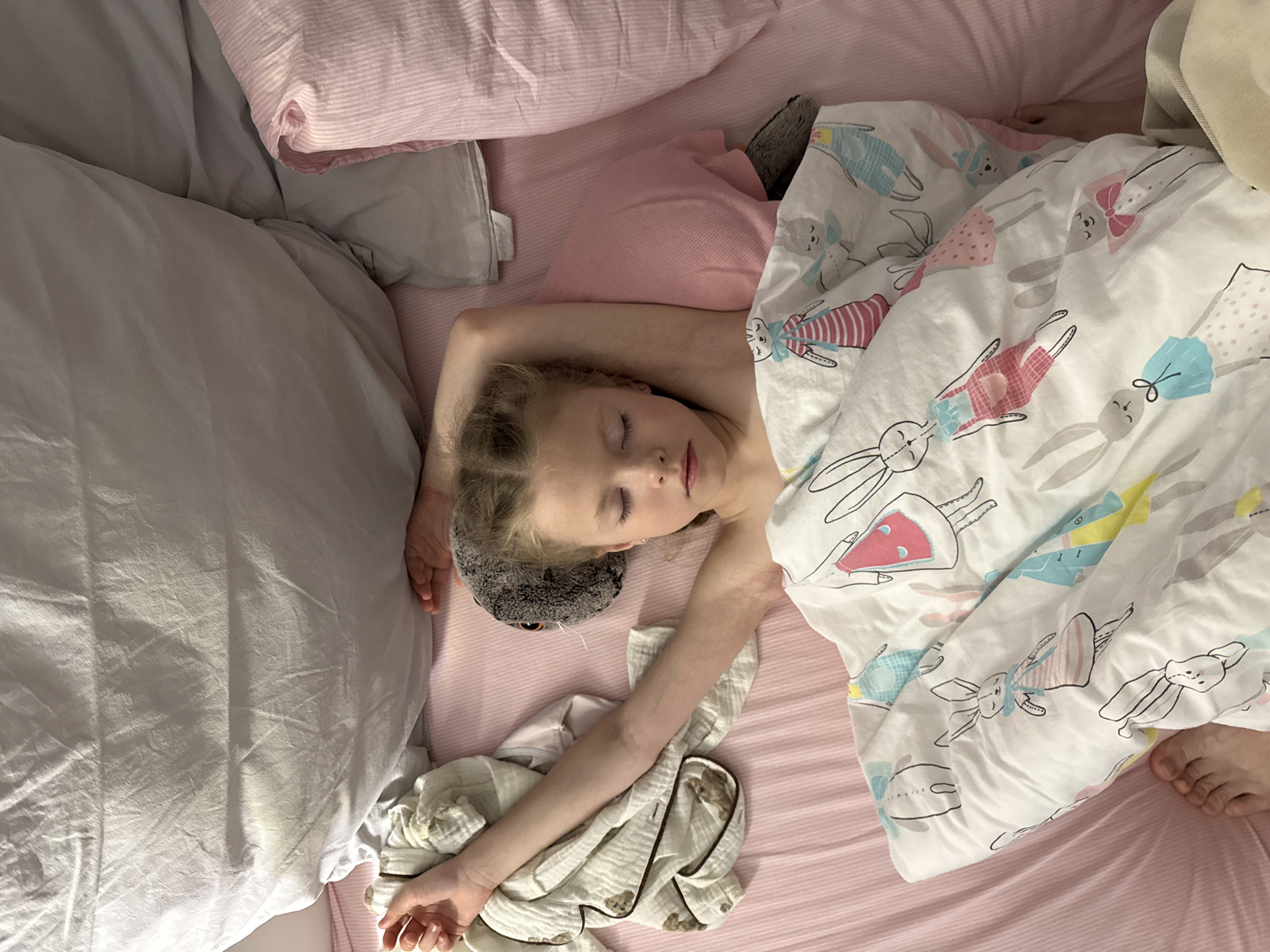 A child is asleep in a bed with light pink sheets and a white comforter patterned with cartoon rabbits. The child lies on their side with their arms extended. A gray stuffed animal is near their head. Pillows of white, pink, and pink with white stripes are also present. The environment is domestic. There is no evidence to suggest a location in Izhevsk, Russia.