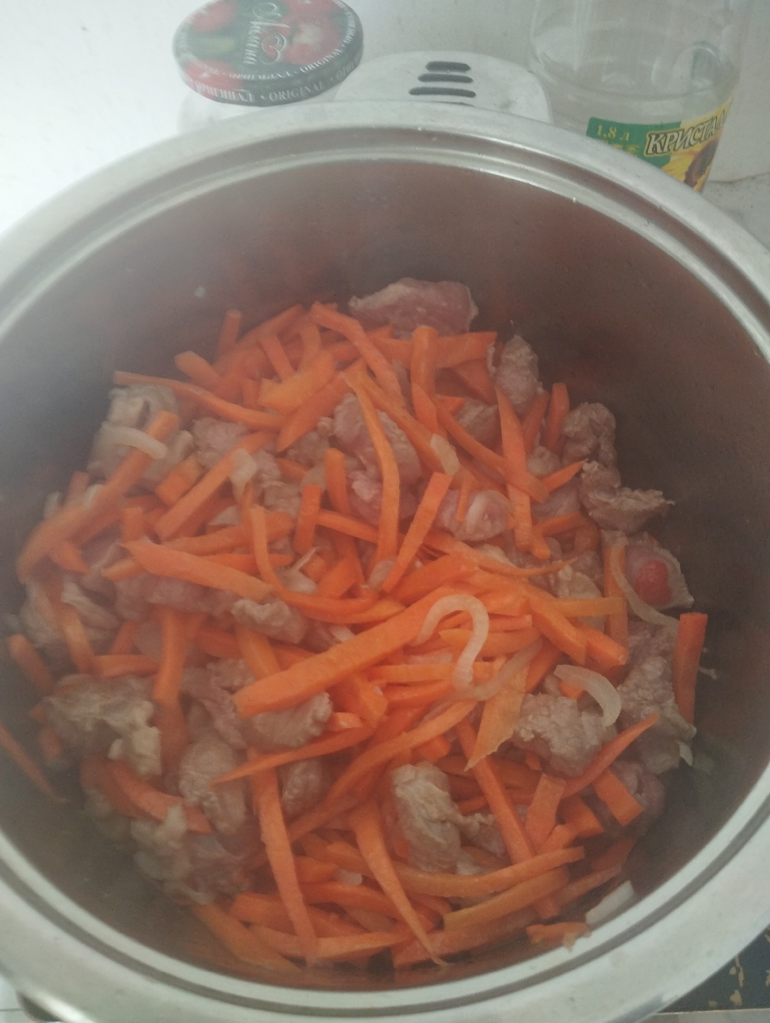 A close-up, overhead shot shows a stainless steel pot filled with sliced carrots and pieces of meat, possibly beef or lamb, suggesting food preparation. The ingredients are roughly chopped and layered within the pot. In the background, out of focus, is a clear plastic bottle with a green label containing Cyrillic text, indicating it's likely a consumer product common in the region. The overall setting appears to be a domestic kitchen environment. The image was taken in Naryn City, Kyrgyzstan.