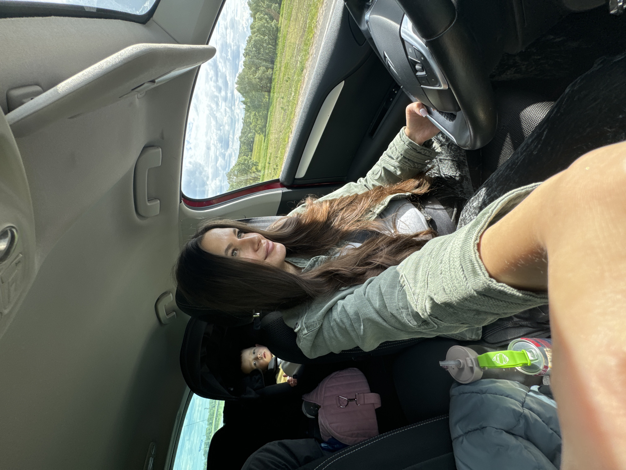 The interior of a car, presumably in motion, reveals a woman in the driver's seat, likely taking a selfie. She has long brown hair and is wearing a light green jacket. She is holding the steering wheel with her left hand. In the back seat, a baby in a car seat is visible, along with a pink bag, a water bottle, and a blue blanket. The car's interior is a mix of black and gray. Through the car window, a green field and some trees can be seen under a cloudy sky. The specific location within the city of Kremyonki, Russia, is not discernible.