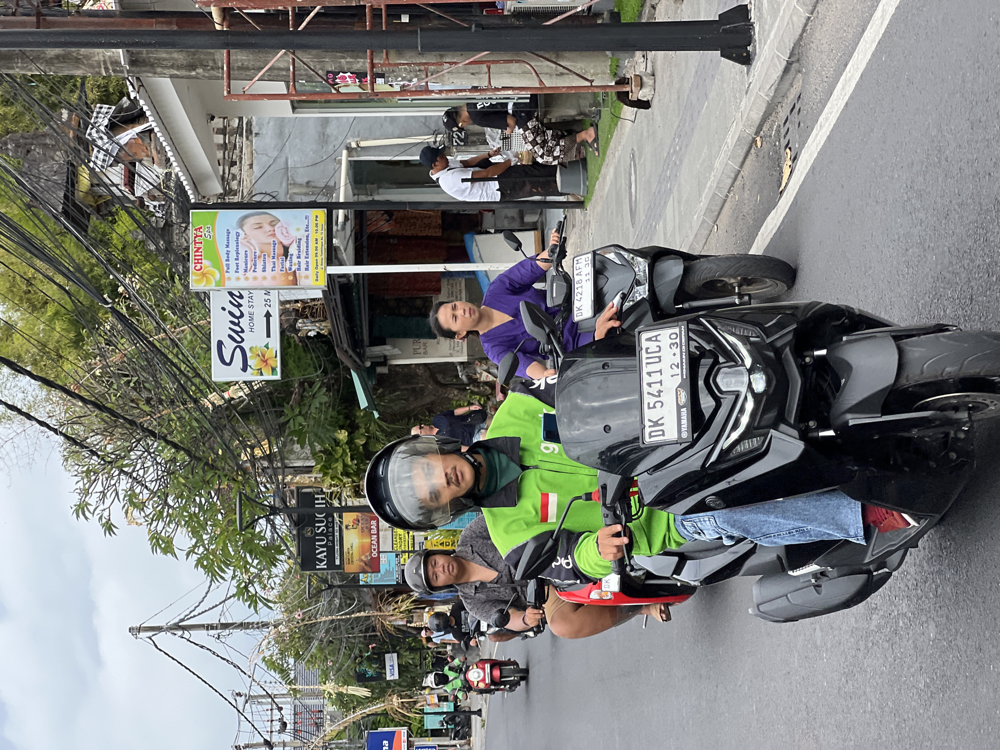 The image depicts a street scene in Denpasar, Indonesia, characterized by a busy road and sidewalk. Several individuals are riding motorbikes, a common mode of transportation in the city. One person in the foreground, wearing a bright green jacket and a helmet, is the primary subject, prominently positioned on a black scooter. Behind them, other individuals on motorbikes are visible, as are pedestrians on the sidewalk. The environment includes buildings lining the street, various signs with text in Indonesian and English advertising local businesses such as "KAYU SUCI PALACE" and "CHINTYA SPA," and abundant overhead electrical wires, typical of urban infrastructure in Bali. The overall impression is one of a bustling, everyday street scene in a tropical urban setting.