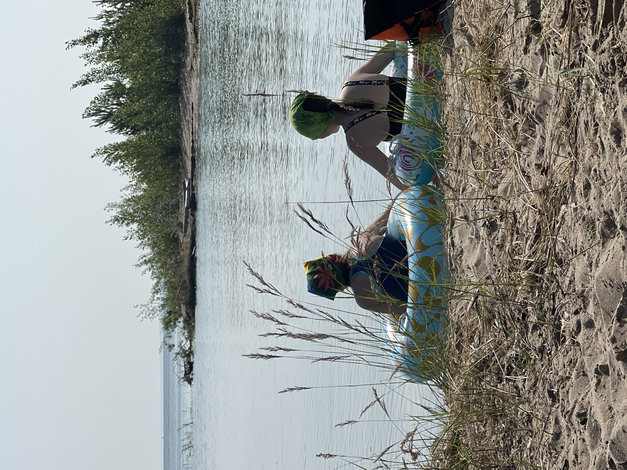 Two individuals, viewed from behind, are seated in inflatable tubes at the edge of a calm body of water. The person on the left wears a multi-colored bandana with a leaf pattern and a blue swimsuit. The person on the right wears a green bandana and a black swimsuit with "NIKE" branding on the straps. Tall, dry grasses in the foreground partially obscure the individuals and their tubes. The water extends to a distant shoreline with a dense line of green trees and bushes. The sky is clear and pale. The overall scene suggests a peaceful outdoor setting by a lake or slow-moving river. The location is not specified as Borovoe, Russia, or any other city.