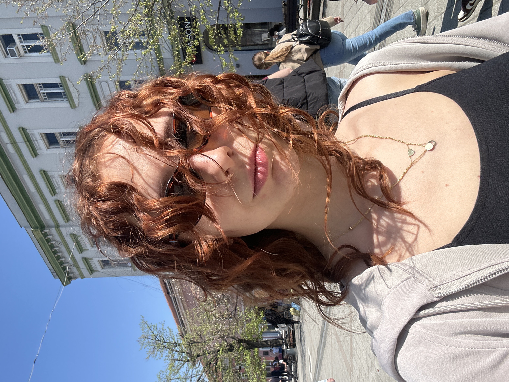 A selfie-style photograph depicts a red-haired woman with sunglasses standing outside on a sunny day. The woman is wearing a black top and a light-colored jacket. She is also wearing a gold necklace.
The background shows a street scene, with buildings, trees, and other people visible. Based on the architecture, the location is most likely in the city of Novi Sad, Serbia. People are walking on the street.