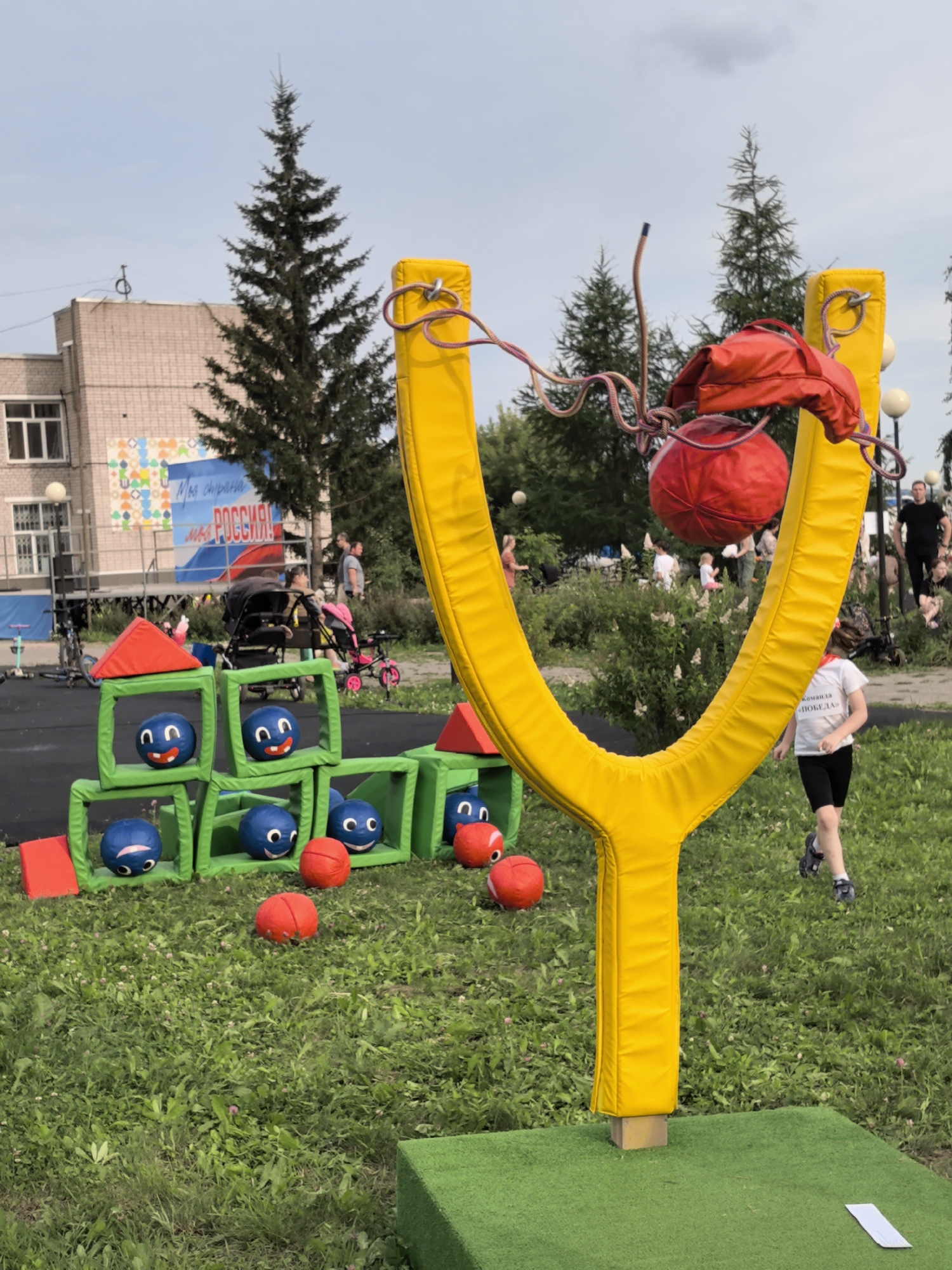A yellow slingshot attraction stands on a grassy area with a building and trees in the background. The slingshot has a red ball suspended from it by ropes. In the foreground, a young boy in a white t-shirt and black shorts runs past the slingshot. To the left of the slingshot, there is a structure made of green blocks with blue balls inside them, each ball having a smiley face. Several red balls are scattered on the grass. In the background, people are visible, and a banner with text that appears to be in Russian is displayed on the building. The overall environment suggests an outdoor event or park in Фролы, Russia.