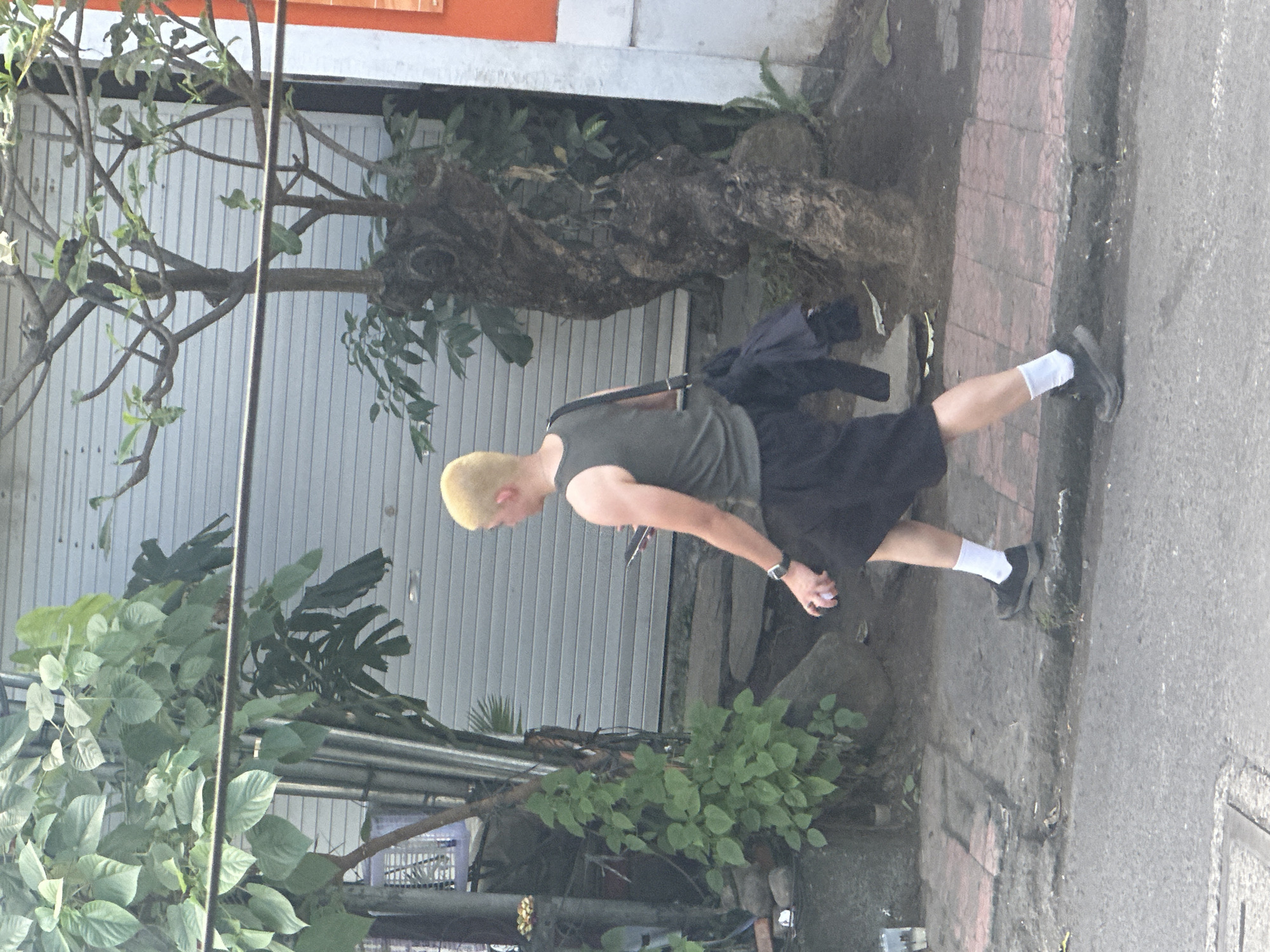 A person with bleached blonde hair is walking on a sidewalk, holding a mobile phone in their right hand. They are wearing a dark green sleeveless shirt, black shorts, white socks, and dark athletic shoes. A dark bag is slung over their left shoulder, with a dark garment draped over it. The sidewalk is paved with reddish-brown bricks and is adjacent to a street. To the left of the sidewalk, there is a large plant with green leaves and a metal pole. Behind the person, a grey roll-up door is visible, and to the right, there is a textured tree trunk. The scene appears to be outdoors in what could be the city of Ubud District, Indonesia, given the tropical foliage and architectural style of the building.