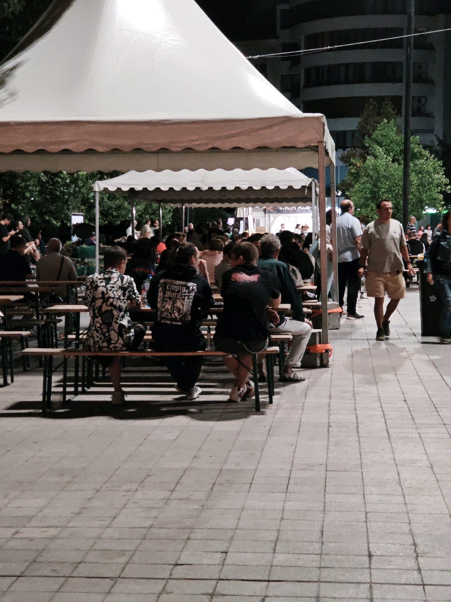 This is a medium shot, captured at night, of a group of people sitting at picnic tables under a large white tent with a scalloped brown and white awning. The tables and benches are made of wood, and the ground is paved with light-colored square tiles. In the background, there are trees, a multi-story building with balconies, and a street-level awning with "STREET FOOD" visible on it.
Several individuals are seated at the tables, facing away from the camera. One person in the foreground is wearing a black hoodie with "Empty Words" written on the back in white lettering. Another person to their left is wearing a black and white patterned shirt. A man in a light-colored t-shirt and shorts is walking away from the tables in the mid-ground. Several other people are visible in the background, some seated, others standing. The lighting is artificial, casting shadows on the tiled ground.