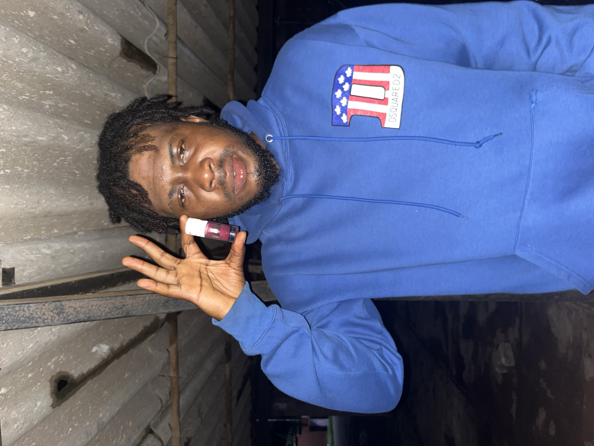 In Jos, Nigeria, a Black man with dreadlocks, wearing a blue Dsquared2 hoodie, stands indoors under a corrugated iron roof with visible water stains. He holds a small, clear bottle containing a pink liquid in his right hand, which is raised to eye level. The man's expression is neutral, and his gaze is directed towards the camera. The background is dark, with hints of outdoor scenery visible.