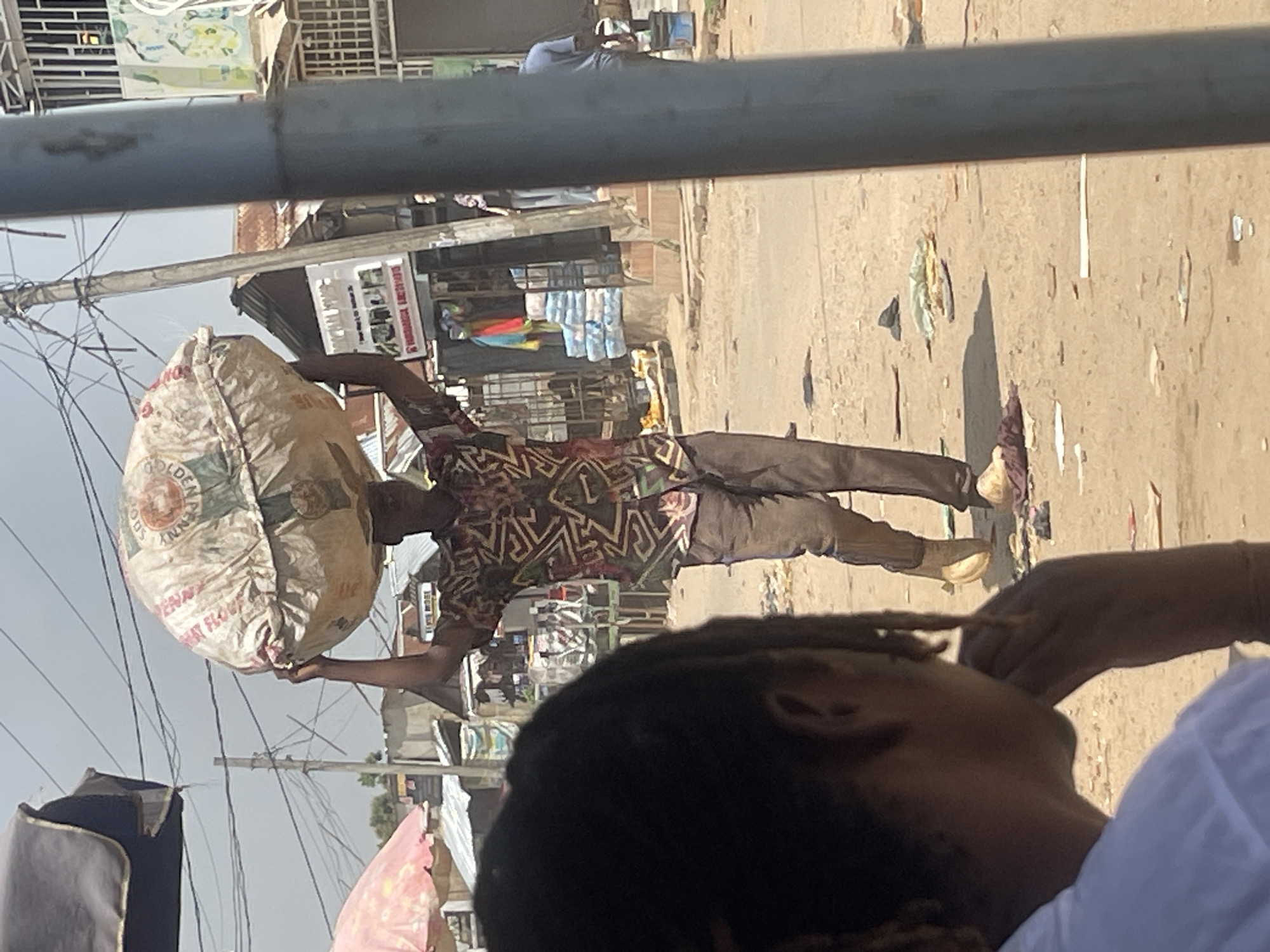 A man carries a large, round, white sack on his head, walking across a dirt path in a bustling outdoor market. The sack appears to be made of woven material and has some text and imagery on it, including what looks like a logo. The man is wearing a brightly colored, patterned shirt and brown trousers. His shoes are visible, and he is mid-stride.
In the foreground, out of focus, is the back of a person with dark, braided hair, wearing a white shirt. This person is looking in the direction of the man with the sack.
The background depicts a typical street scene in Abuja, Nigeria, with market stalls, buildings, and other people. There are several structures with corrugated metal roofs, some with awnings providing shade. Colorful fabrics and stacks of goods are visible in the market stalls. Overhead, power lines and poles crisscross the sky. The ground is a mixture of dirt and debris, suggesting a well-trafficked area. The lighting indicates it is daytime, with bright sunlight casting shadows. The overall impression is of a vibrant and active market environment.