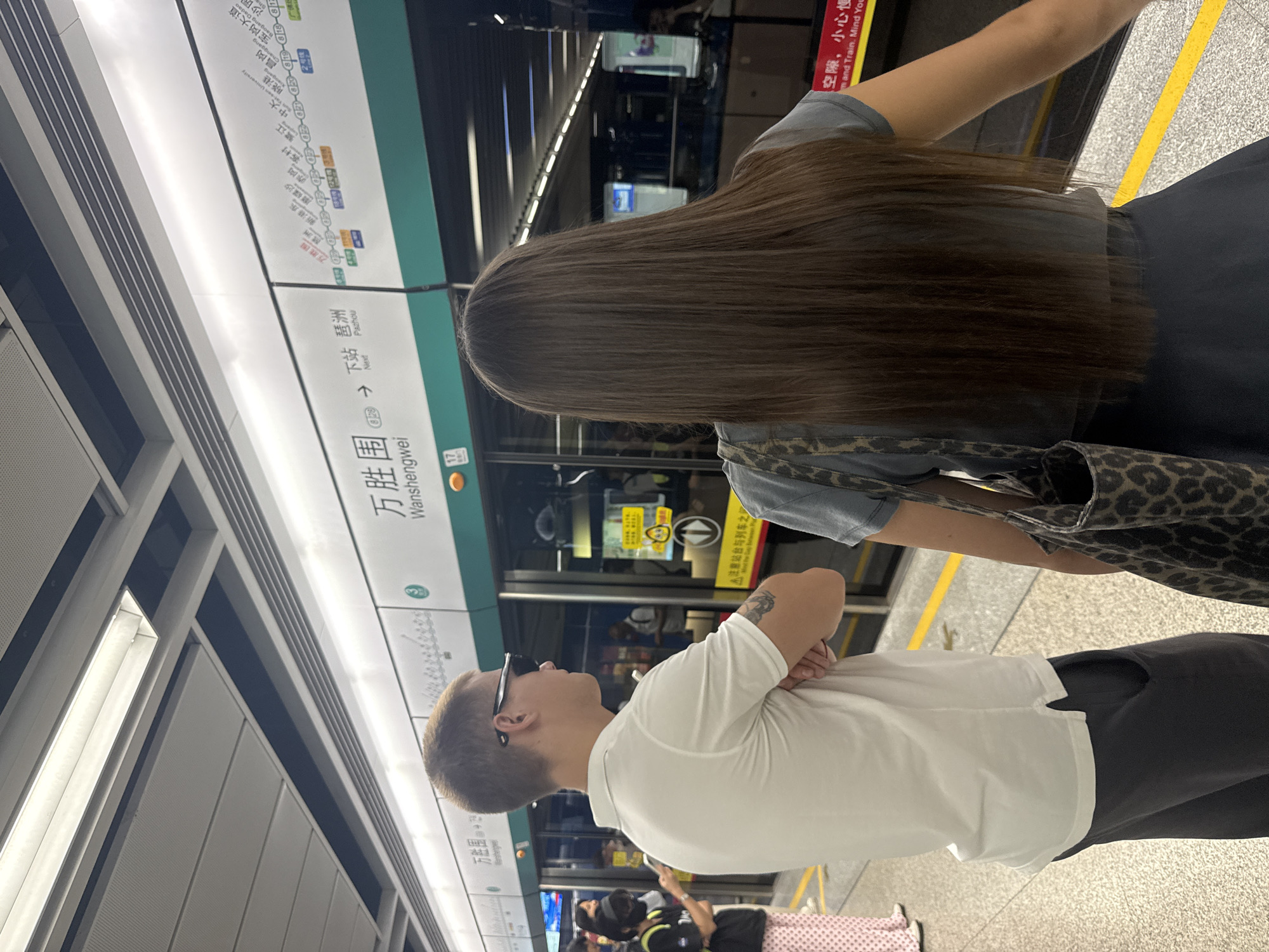 This is a medium shot, taken at a slight upward angle, of two individuals waiting on a subway platform in Haizhu District, China. The platform is clean and well-lit, with a yellow safety line marking the edge. Behind the individuals are the closed doors of a subway train, featuring a route map with Chinese characters and English station names, indicating the line serves "Wanshengwei" and "Pazhou." A man wearing a white polo shirt and dark pants stands on the left, with his arms crossed and wearing sunglasses. A woman with long brown hair, wearing a gray t-shirt and a dark skirt, stands on the right, carrying a leopard-print tote bag. Other passengers are visible in the background, also waiting for the train. The overall scene depicts a typical urban subway waiting area.