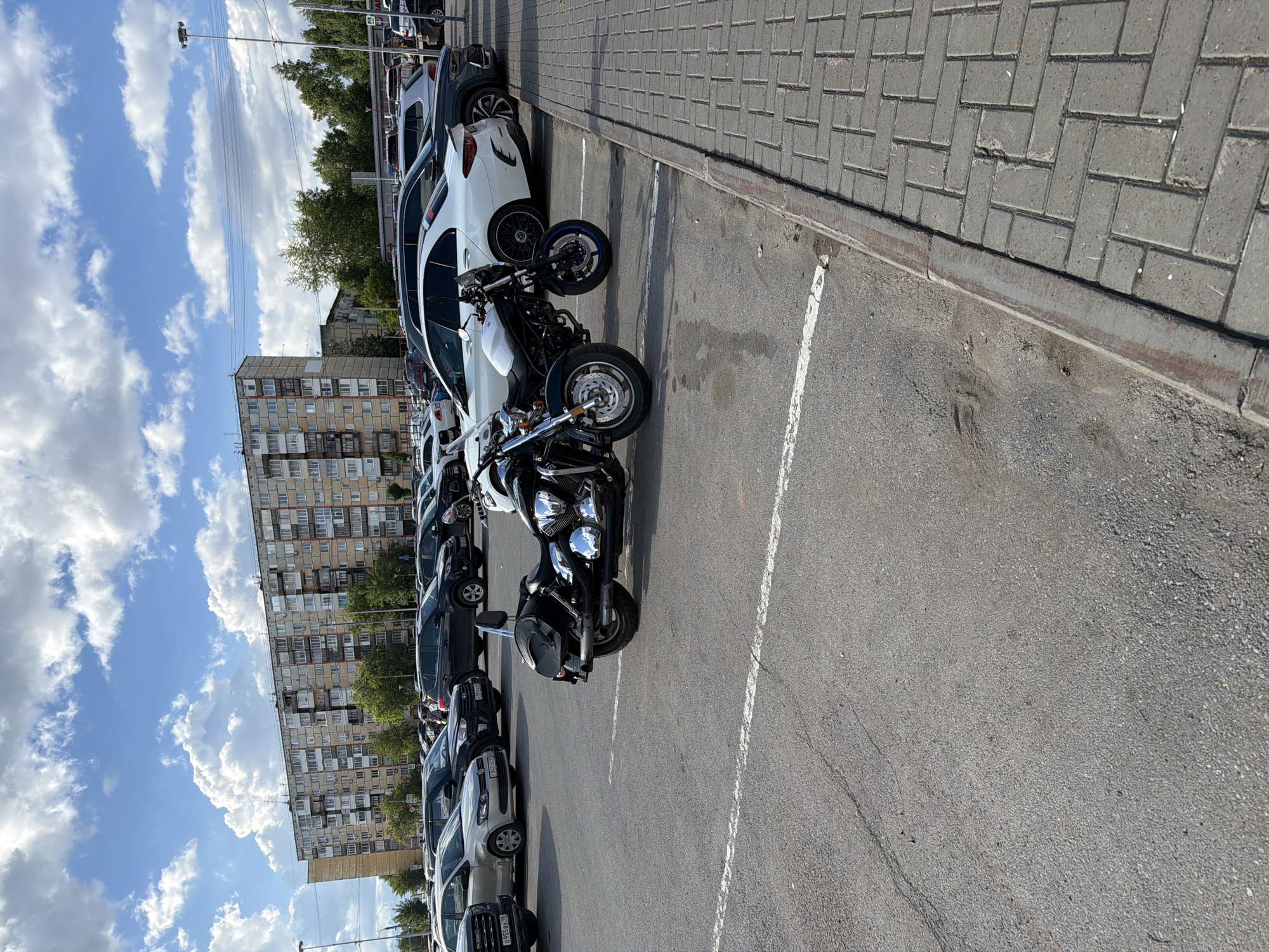 The image depicts a parking lot scene in Izhevsk, Russia. Several vehicles, including cars and motorcycles, are parked in marked spaces. A tall apartment building stands in the background. The sky is partly cloudy. The parking lot is paved and bordered by a brick walkway.