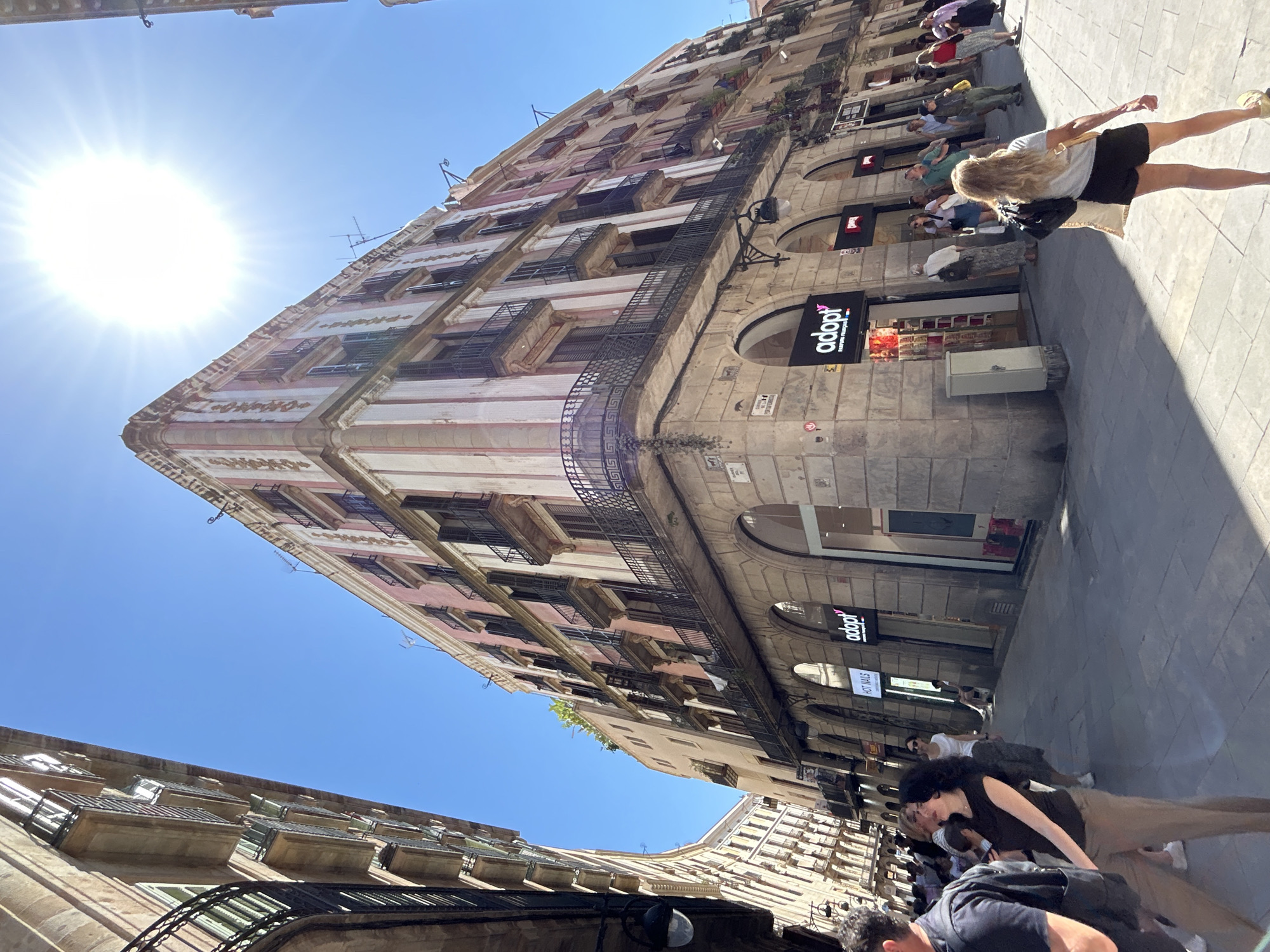 A street scene in Barcelona, Spain, is captured from a low angle, looking up at a tall building with a pink and white facade. The building has multiple floors, each featuring small balconies with dark metal railings. On the ground level, there's a shop with a sign that reads "adopt". People are walking along the sidewalk in front of the building, including a woman with long blonde hair wearing a black skirt. The sky is clear and blue, and the sun is shining brightly, creating a strong glare. Another building is visible on the lower left side of the frame, also with balconies and architectural details. The street appears narrow, with other pedestrians and details of buildings extending into the background, hinting at a bustling urban environment.