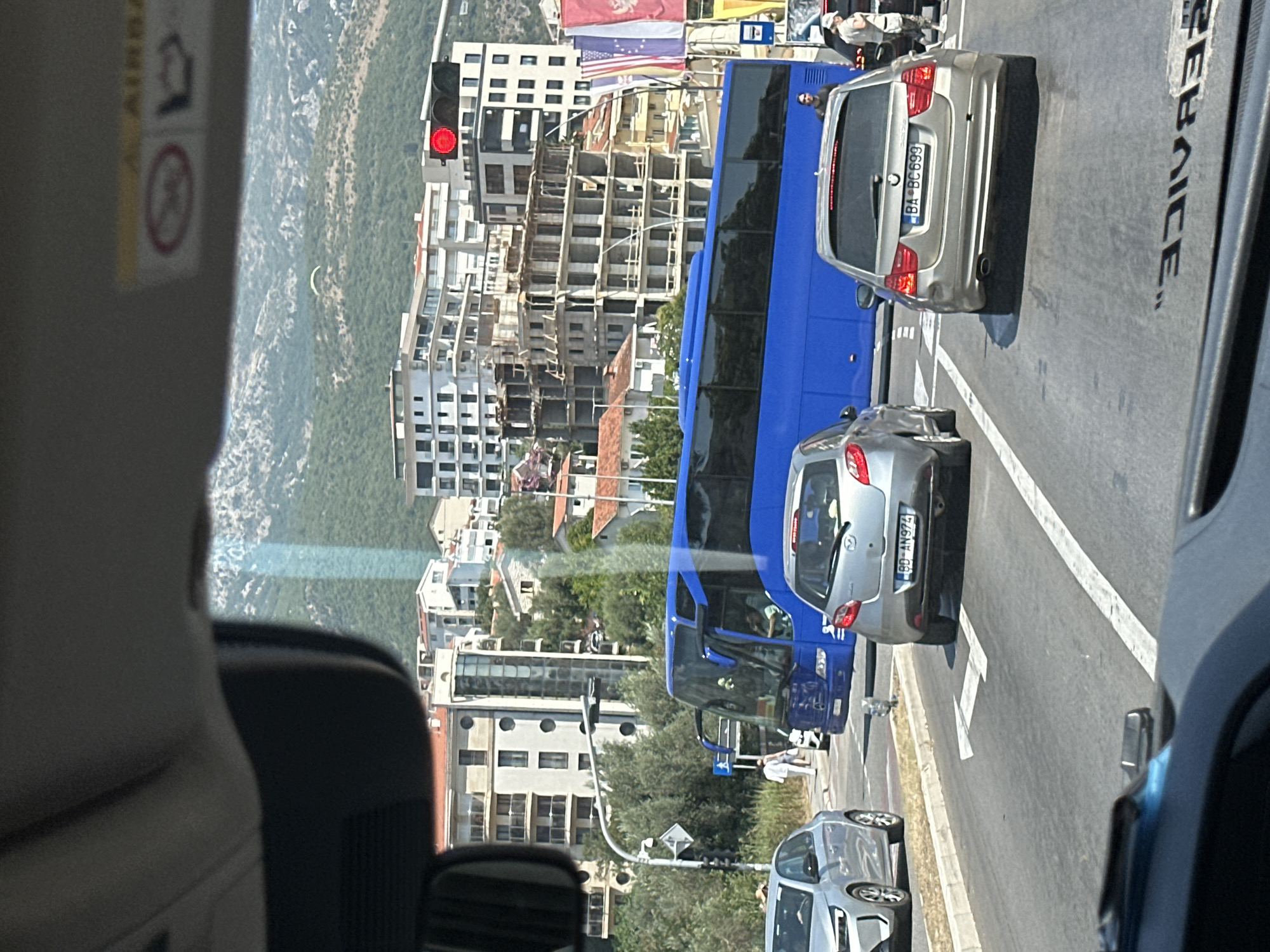 The image is taken from inside a vehicle, looking forward through the windshield. It depicts a street scene in Boreti, Montenegro, during the daytime. The foreground shows the interior of the vehicle, with a rearview mirror on the left and the dashboard visible at the bottom.
The main subjects in the image are several vehicles on a paved road. A large blue bus is prominent, positioned centrally. In front of the bus, a silver hatchback car with license plate "BD-AN974" is stopped. To the right of the hatchback, a silver BMW wagon with license plate "BA-BC 699" is also stopped. To the left of the bus, a silver sedan is visible, partially obscured. There are also pedestrians on the sidewalk in the background, including one person in a white shirt walking near the bus.
The environment consists of a street with lane markings. In the background, there are multi-story buildings with modern architecture, some of which appear to be under construction. Beyond the buildings, a lush green hillside with rocky outcrops rises, and a paraglider is visible in the sky above the mountains. A traffic light with a red signal is mounted on a pole to the right of the scene. Several national flags, including what appears to be the flag of Montenegro, are displayed on poles to the right of the bus. The overall impression is a busy urban environment with a backdrop of natural mountainous landscape.