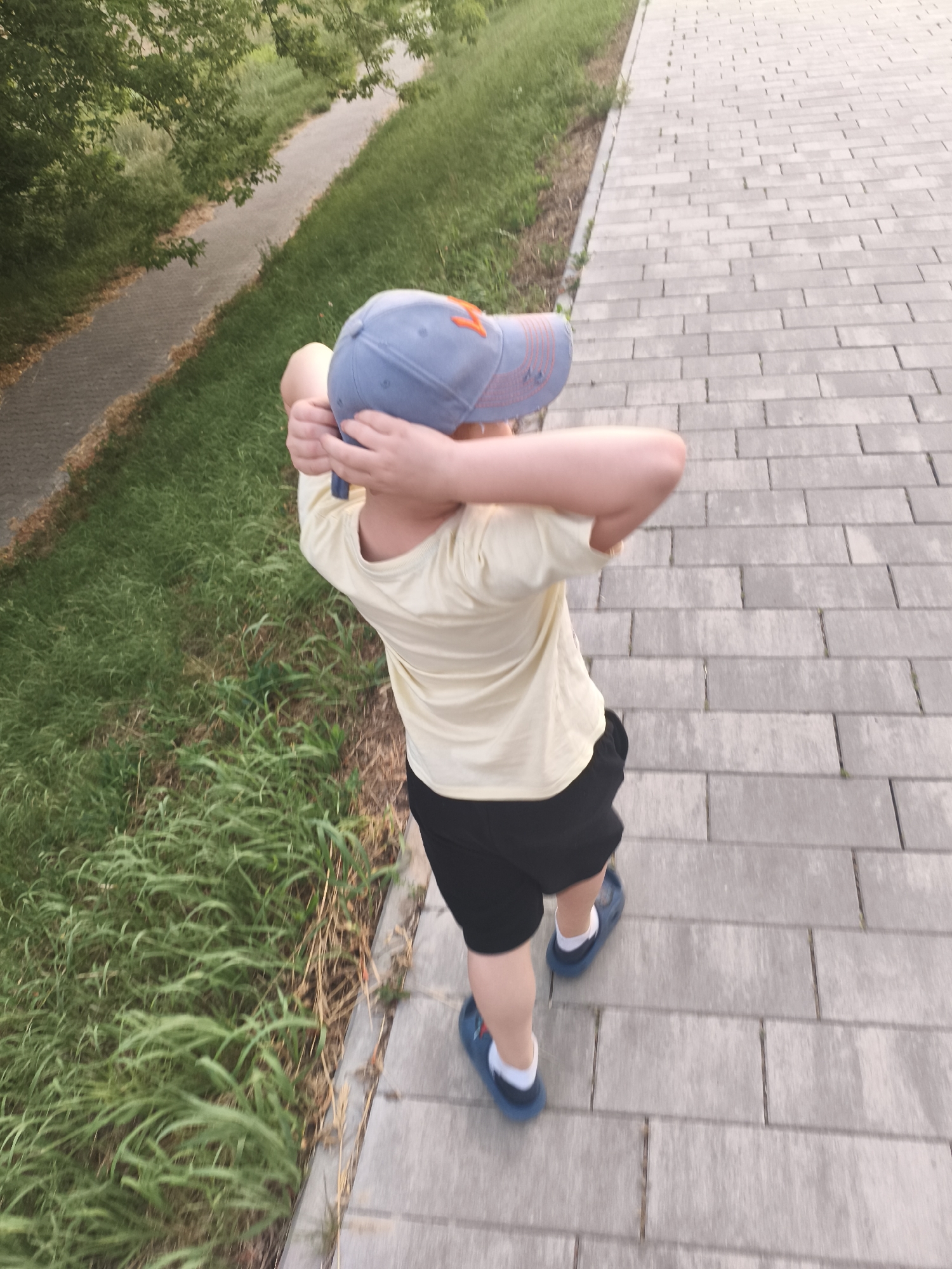 A young person, seen from a high angle, is walking on a paved path. They are wearing a pale yellow t-shirt, black shorts, and blue clogs with white socks. They have a blue baseball cap on their head, which they are holding onto with both hands, obscuring their face. The cap has an orange emblem on the front. The path is made of rectangular gray pavers and runs alongside a grassy area. Beyond the grass, a paved walkway is visible, bordered by trees and more grass. The scene appears to be outdoors during the daytime, likely in the late afternoon or early evening, given the lighting. The overall mood is casual and relaxed, with the child seemingly enjoying a walk. There is no visible text in the image. The location is identified as Nowy Dwór Mazowiecki, Poland.