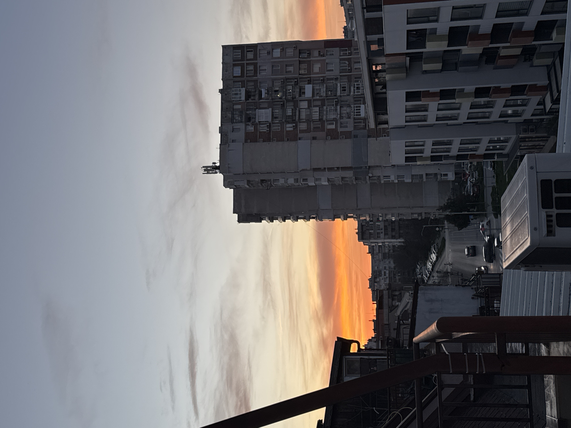 The image depicts a sunset view in a city setting, likely Varna, Bulgaria, given the architecture and urban layout. The foreground shows rooftops and metal structures, suggesting the photograph was taken from an elevated position. Mid-ground features tall, Soviet-era apartment buildings with numerous windows and balconies, common in Eastern European urban areas. A street runs between buildings, with cars parked along the sides. The sky exhibits a gradient of colors, with orange and yellow hues near the horizon, indicative of a sunset. The overall atmosphere suggests an urban landscape at dusk.
