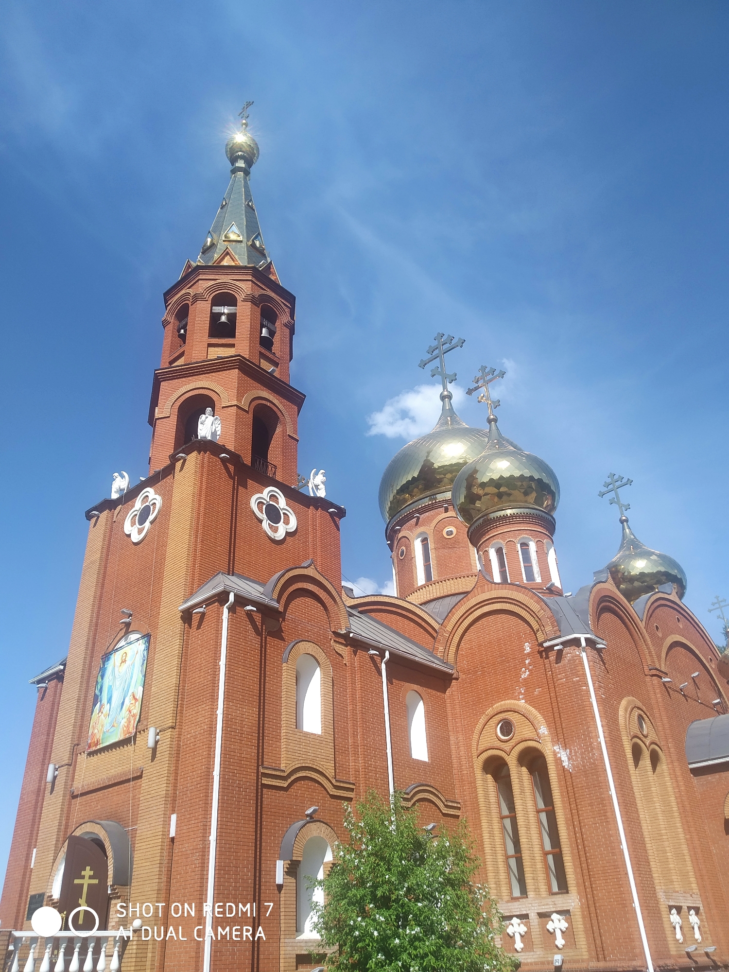 The image shows the Cathedral of St. George the Victorious, located in Chaikovsky, Russia. It is a brick building with a tall bell tower and golden domes topped with crosses. The building also features decorative elements like white angels and a large image of a saint. The sky is blue with scattered clouds.