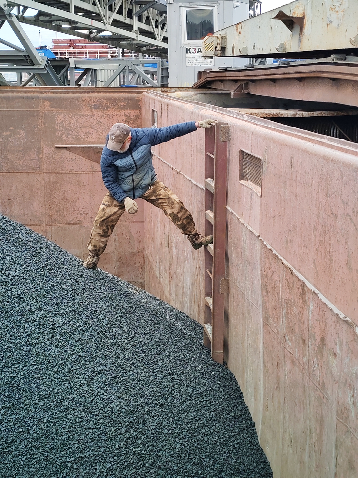 A man is engaged in activity within a large, weathered, reddish-brown metal cargo hold or container, likely at an industrial port in Shcheleiki, Russia. He wears a blue puffer jacket, camouflage-patterned pants, a brown baseball cap, and light-colored work gloves, and has a visible beard.
The man is positioned on a steep pile of dark grey aggregate, such as gravel or crushed stone, which fills a significant portion of the hold. His left foot is on the aggregate, while his right foot is on a rung of a sturdy metal ladder attached to the inner wall of the container. He is using his right hand to grip the top edge of the wall for balance, indicating he is either climbing into or out of the hold, or inspecting the cargo.
In the background, industrial infrastructure is visible. Large, grey metal beams and supports, characteristic of a gantry crane or a bulk material handling system, dominate the upper portion of the image. A white portable cabin or control room stands behind these structures. On the cabin, the prominent text "K3A" is visible, along with smaller, partially legible contact details, including a German phone number (+49). Further in the distance, a red ship or another part of a port facility can be seen. Text on the red structure includes "ГРАВИТА ПОДЪЕМОМ" (GRAVITA POD'YOMOM), "БООВ", and "200В".
The time of day appears to be daytime, under an overcast sky, as indicated by the diffused lighting and lack of harsh shadows. The overall scene depicts a laborious task within a working industrial environment.