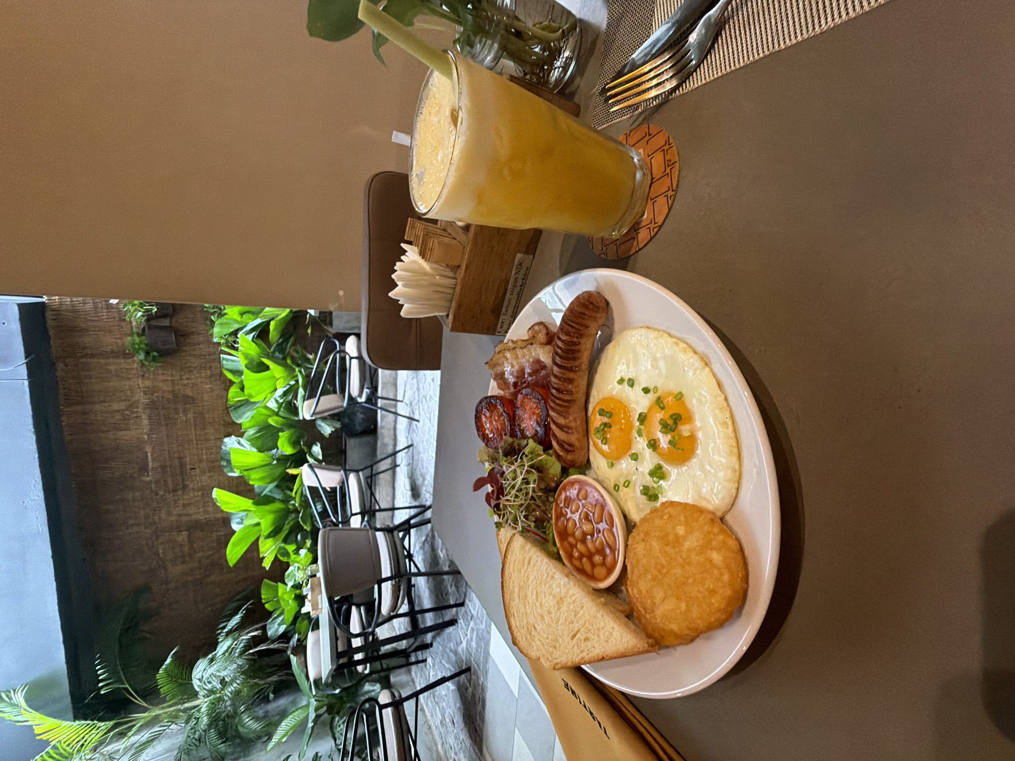 The image displays a detailed view of a breakfast or brunch setting at an indoor-outdoor cafe, specifically identified as Trattoria Saigon NDC in Thủ Đức, Vietnam, based on legible text on a menu folder.
In the immediate foreground, a dark grey tabletop holds a white oval plate featuring a full breakfast meal. This meal comprises two slices of toasted white bread, a small ceramic ramekin containing baked beans in tomato sauce, a round golden-brown hash brown patty, two fried eggs garnished with chopped green onions, a portion of grilled halved tomatoes, several strips of crispy bacon, a grilled sausage, and a small side salad composed of mixed greens and sprouts.
To the right of the plate, a tall glass filled with a frothy, yellow beverage, likely juice, containing ice, rests on a circular coaster with a geometric pattern. A light green stalk, possibly lemongrass or sugarcane, serves as a straw. Further right, a set of silver cutlery (fork and knife) is positioned on a striped placemat.
Behind the plate, a wooden caddy holds a stack of white folded napkins and individual sachets, possibly sugar or condiments. Adjacent to the caddy, a small glass jar contains a green plant cutting submerged in water.
The background reveals a cafe environment designed with natural elements. Lush green foliage, including palm fronds and large-leafed plants, fills much of the middle ground. Behind the plants, a dark wooden slatted wall or fence provides a rustic backdrop. Multiple vacant cafe tables with dark tops and chairs with black metal frames and light-colored padded seats are visible across a light-colored, patterned stone or tiled floor, suggesting an open-air or semi-covered dining area. No individuals are present within the frame.
