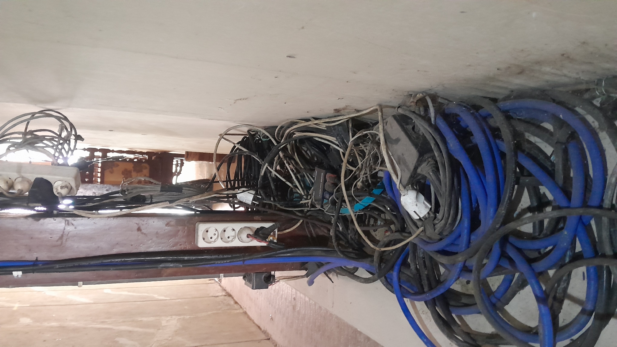 A chaotic tangle of electrical cords and extension cords is piled on the floor and against a wall, obscuring a doorway or narrow passage. The cords are of various colors and thicknesses, including prominent blue ones. Several power outlets are visible, including a multi-socket strip attached to a wooden pole and two individual wall sockets. Some plugs are inserted into these outlets. The environment appears to be indoors, with textured walls and a wooden structural element. The overall impression is one of disorganization and potential electrical hazard. No individuals are present in the image. The scene is likely located in Blora, Indonesia, based on the prompt.