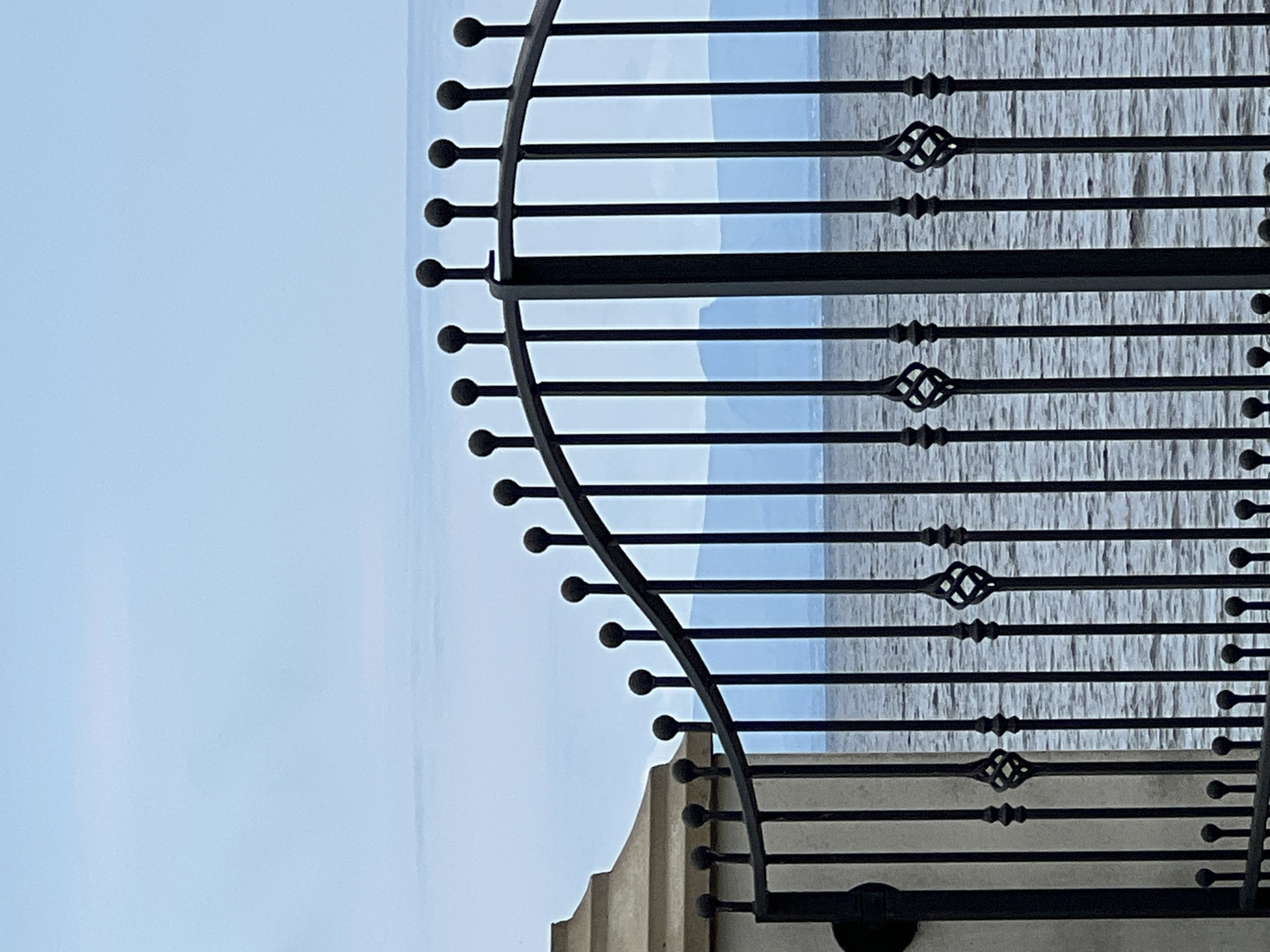 The image shows a black wrought iron gate in front of a body of water. Beyond the water, faint mountain ranges are visible against a pale blue sky. To the left of the gate, a portion of a stone pillar is visible. The gate has decorative finials on top of each vertical bar and twisted decorative elements between some of the bars. The water appears choppy. This scene is likely located near the coast in the town of Mountcharles, Ireland. There are no people or animals present in the image, nor any discernible actions or interactions.