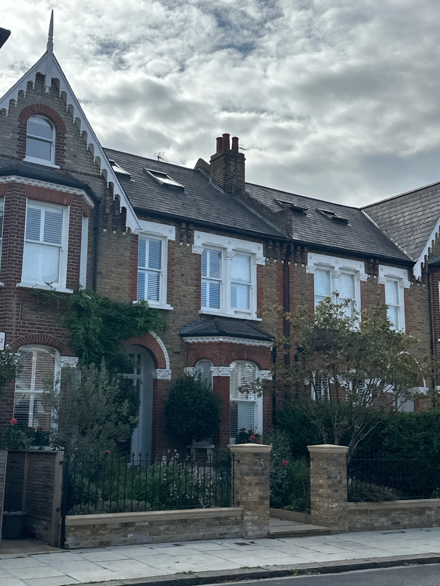 The image shows a row of terraced houses in London, United Kingdom. The houses are built of brick and have pitched roofs with dormer windows. The windows on the ground floor and first floor are sash windows with white shutters. The houses are fronted by a garden with a black metal fence and a low brick wall. The number "25" is visible on a brick pillar at the entrance to the garden. The sky is overcast with clouds.