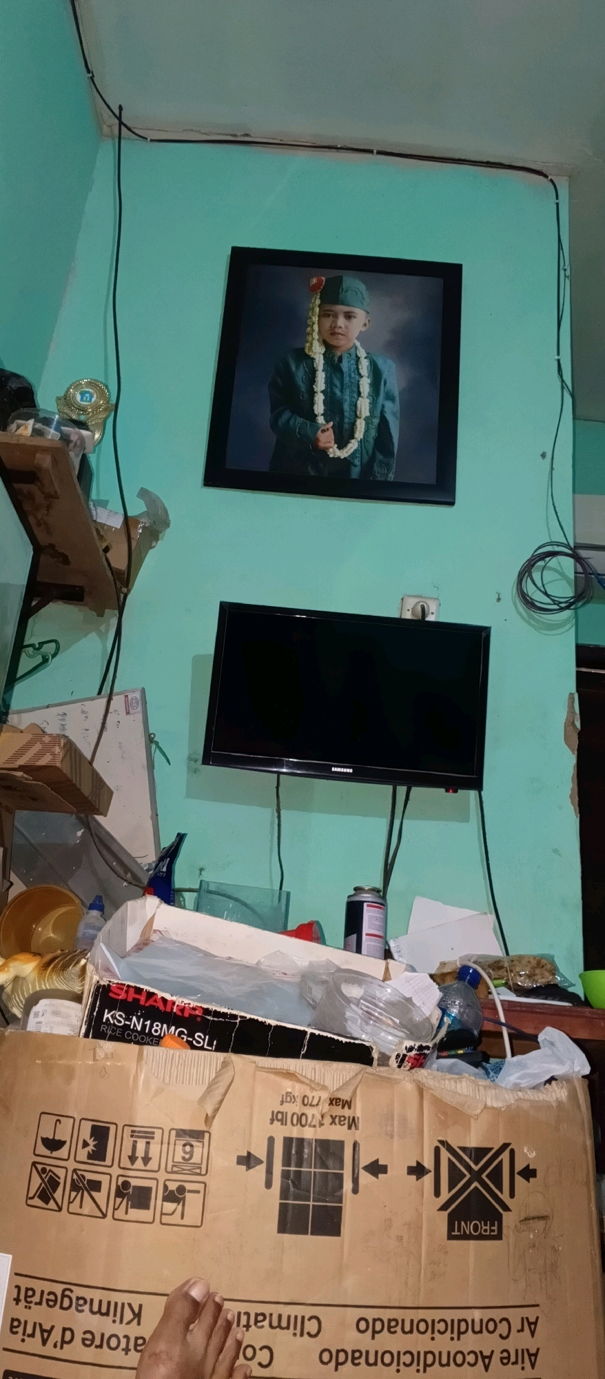 The image shows a wall in a room. A framed portrait of a young boy wearing traditional attire is hung on the wall. Below the portrait, a black flat-screen television is mounted. To the left of the television, there are shelves with various items, including a trophy, boxes, and a plastic bowl. Below the shelves and to the left of the television, there is a large cardboard box with shipping labels and symbols. A person's foot is visible in the lower left corner of the image, resting on the cardboard box. The room appears cluttered with objects and cables are visible along the wall and near the television. The wall color is light blue-green. The location is identified as Watubelah, Indonesia, though the image itself does not provide direct visual confirmation of this specific location beyond the text on the box.