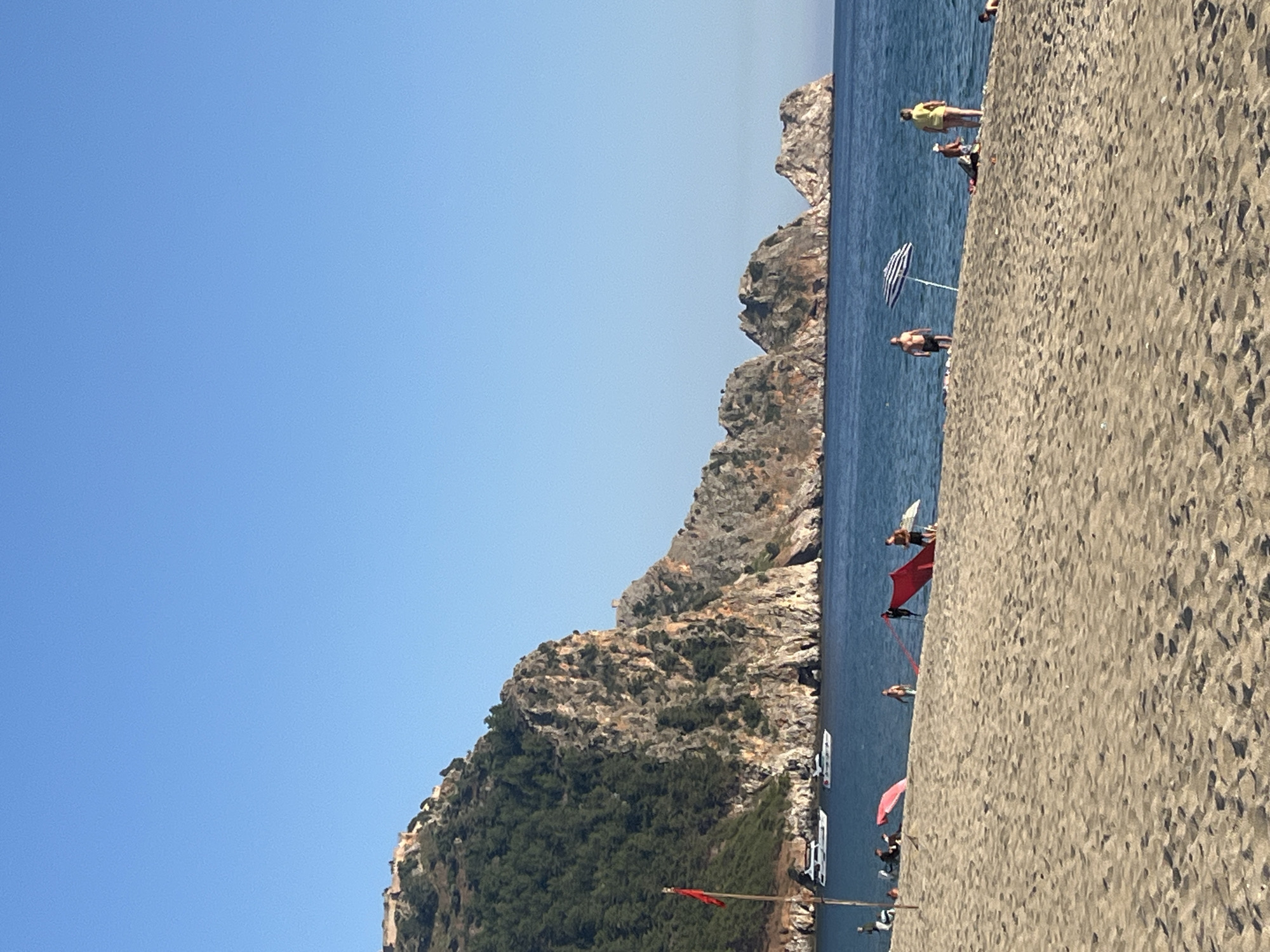 This is an outdoor shot of a beach in Alanya, Turkey. The beach is situated along the coast with cliffs or rocky hills covered in greenery rising on one side. The water is blue and calm, and people are seen swimming, sunbathing, and relaxing on the beach. There are boats anchored close to the shore, and umbrellas provide shade for beachgoers. The sand is light in color and appears coarse. The sky is clear and blue.