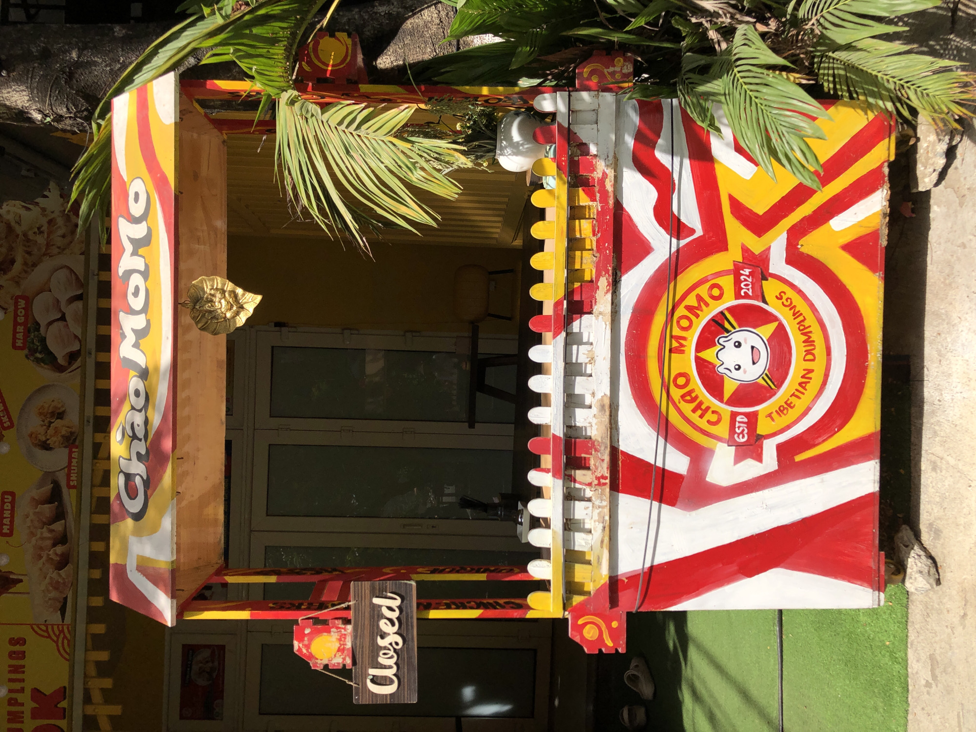 A bright, colorful food stall named "Chao Momo" is set up outdoors in Đà Nẵng, Vietnam. The stall is predominantly red, yellow, and white, with bold graphic designs. A "Closed" sign hangs from a wooden post on the left side of the stall. Above the stall, a yellow banner displays images of dumplings with text labels like "MANDU," "SHUMAI," and "HAR GOW." A golden leaf-shaped ornament hangs from the stall's awning. The stall is situated on a green, carpet-like surface, with a concrete walkway visible in the foreground. Lush green palm fronds and other plants frame the stall on the right side. The background shows a building with glass doors and yellow walls. There are no people visible in the image.