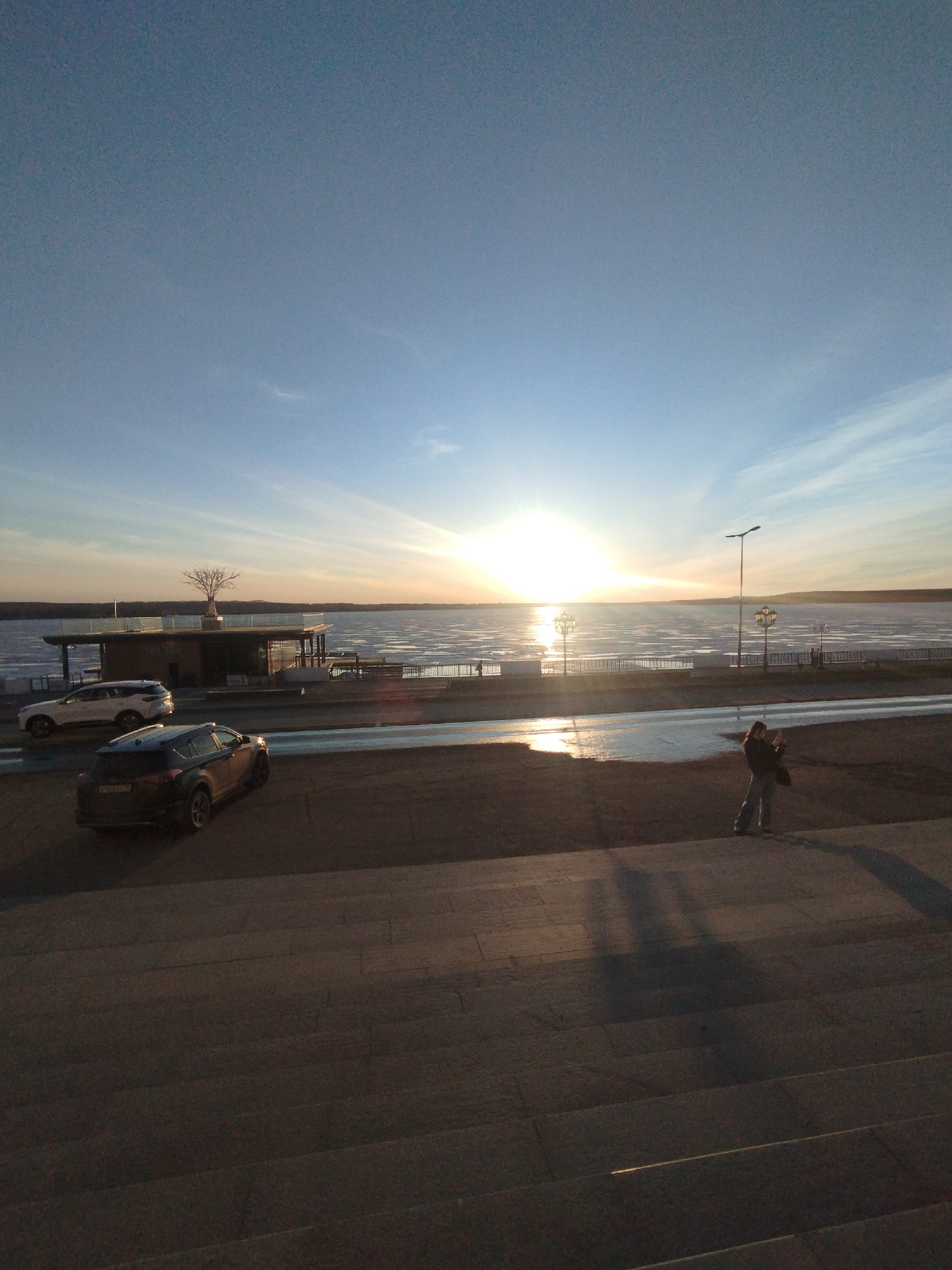 The image shows a sunset scene at a waterfront area in Votkinsk, Russia. A frozen body of water stretches across the horizon, reflecting the setting sun. A building with a tree on top and some parked cars are visible on the left. A person stands on a paved area, possibly taking a picture of the landscape. The foreground consists of steps leading down to the area. Light posts are visible, and the overall lighting indicates it is either early morning or late afternoon.