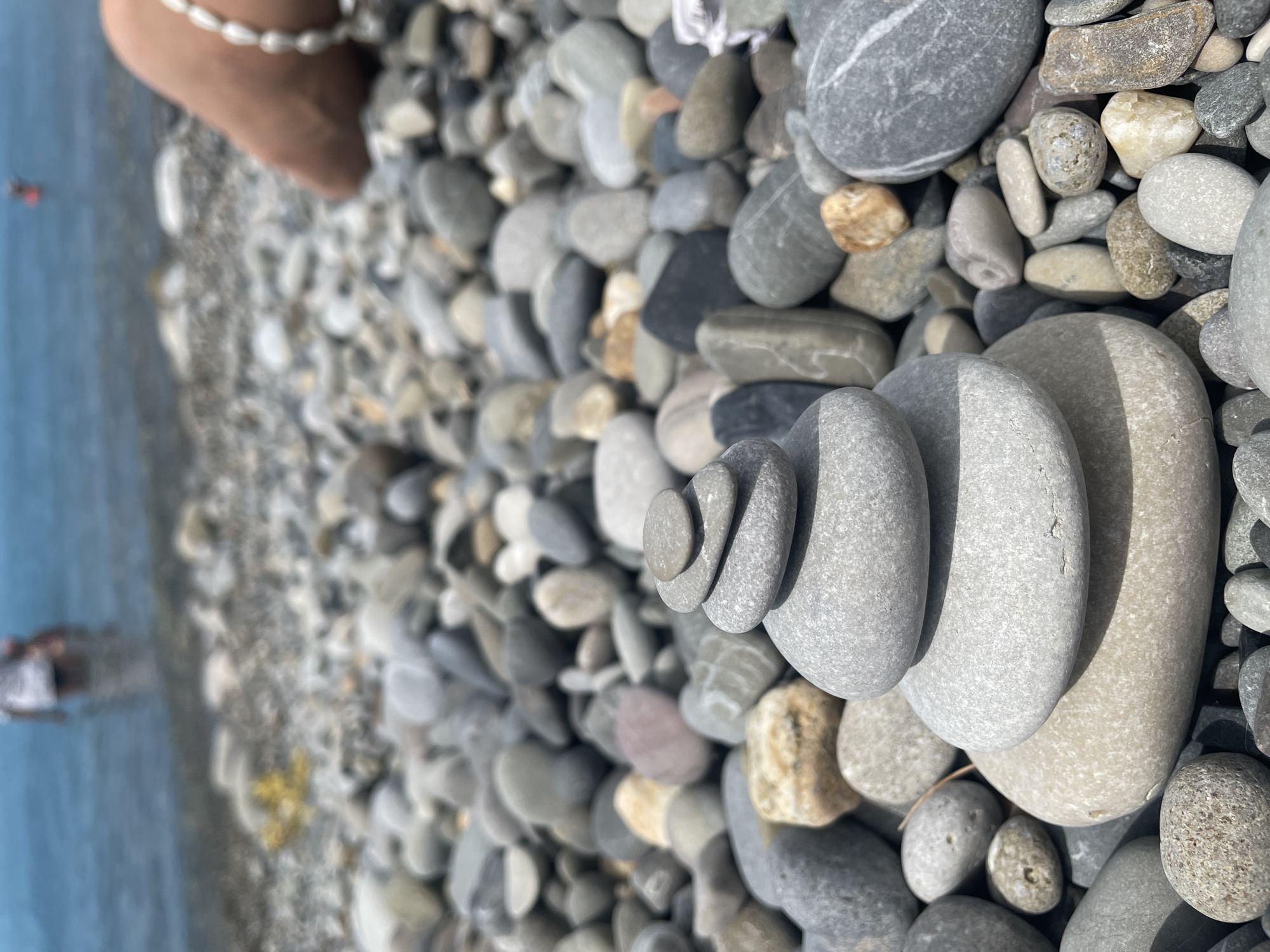 A low-angle, eye-level shot captures a stack of five smooth, grey and tan pebbles on a pebble-strewn beach. The pebbles are stacked in decreasing size from bottom to top, with the largest at the base being a tan color and the smallest at the top being a light gray. Behind the stacked pebbles, the beach continues with a dense scattering of various-sized grey, tan, and off-white pebbles. In the background, a person stands in shallow, blue water, partially obscured and out of focus. The horizon line is visible where the water meets the sky, which is a pale blue. To the right of the frame, a person's leg and foot are visible, adorned with a pearl bracelet. The lighting suggests a sunny day, casting subtle shadows on the pebbles.