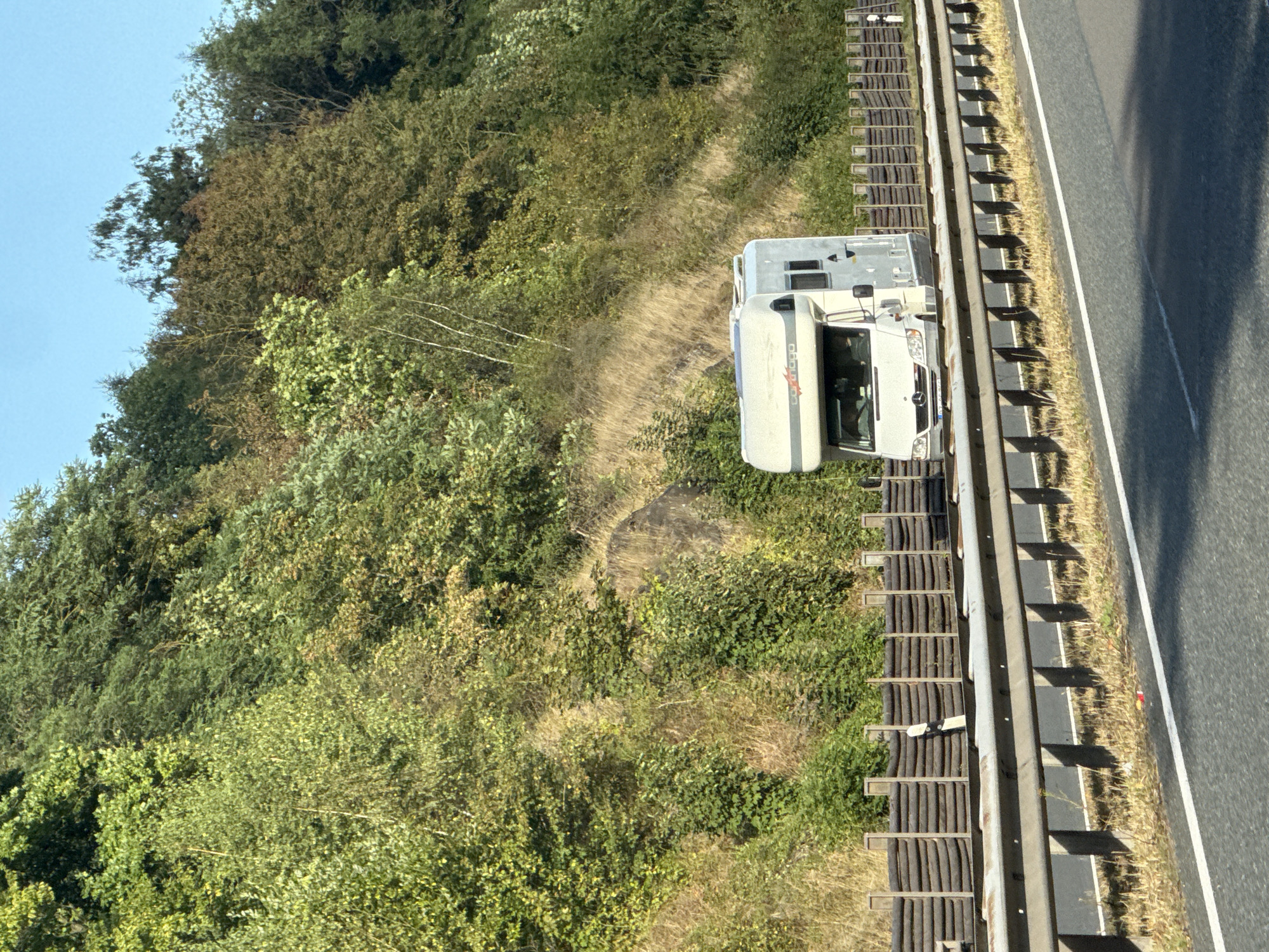 The image shows a white recreational vehicle (RV) driving on a highway. The RV is positioned on the right side of the road, partially obscured by a guardrail and a sound barrier. The RV has a visible logo on its side that reads "CARTHAGO." The background consists of a densely vegetated hillside with various shades of green foliage. The sky is clear and blue. The foreground displays a section of the highway with lane markings and a guardrail. The location is identified as Herzberg, Germany.