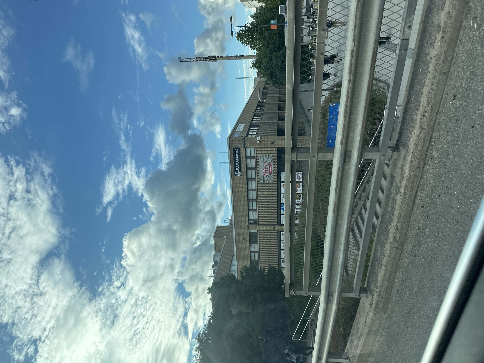 The image shows a building with a sign that says "MAMA BOUTIQUE". There are also signs for "Mero MARKET". A tall structure with antennas or similar equipment on top is visible in the background, with the word "STADION" printed on it. The scene appears to be a street or an area near a stadium. The sky is blue with scattered clouds, suggesting it's daytime and the weather is partly cloudy. A metal railing runs along the foreground, which is likely a bridge or walkway overlooking the street and building. There are some blurred figures of people visible behind the railing. The building is made of brick and has multiple windows.