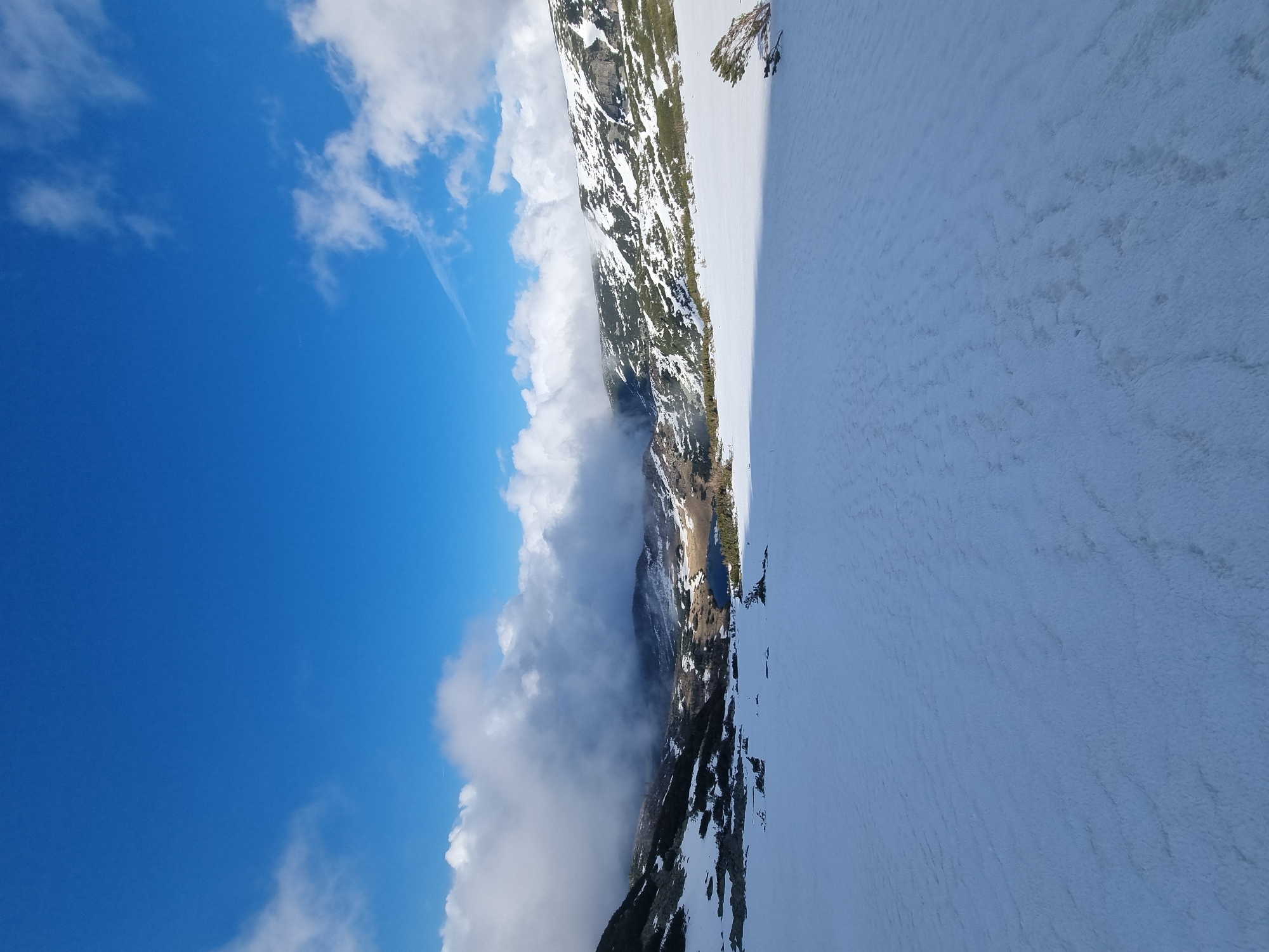 The image shows a mountain landscape near Govedartsi, Bulgaria. A mountain ridge partially covered in snow and some sparse vegetation separates a valley filled with cloud cover on the left from a large snow-covered surface on the right. The sky is a clear blue with scattered white clouds.