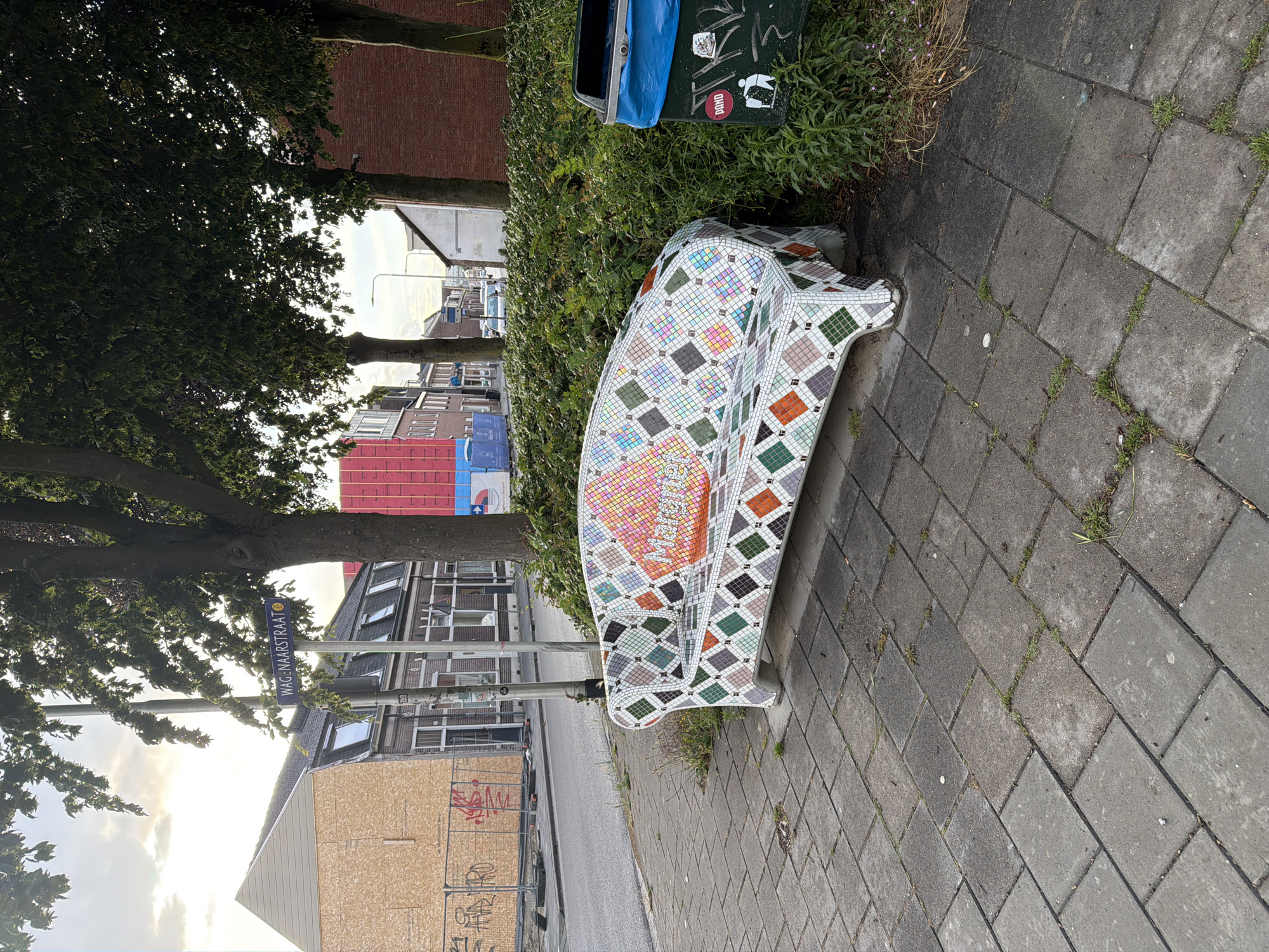 A mosaic-covered bench sits on a tiled sidewalk next to a trash can and lush green bushes. The bench is decorated with a diamond pattern in various colors, with the name "Marmette" visible on its surface. In the background, a street is lined with buildings, one of which is under construction with plywood covering its facade. A street sign reads "WAGENAARSTRAAT". The scene is set outdoors in a city environment.