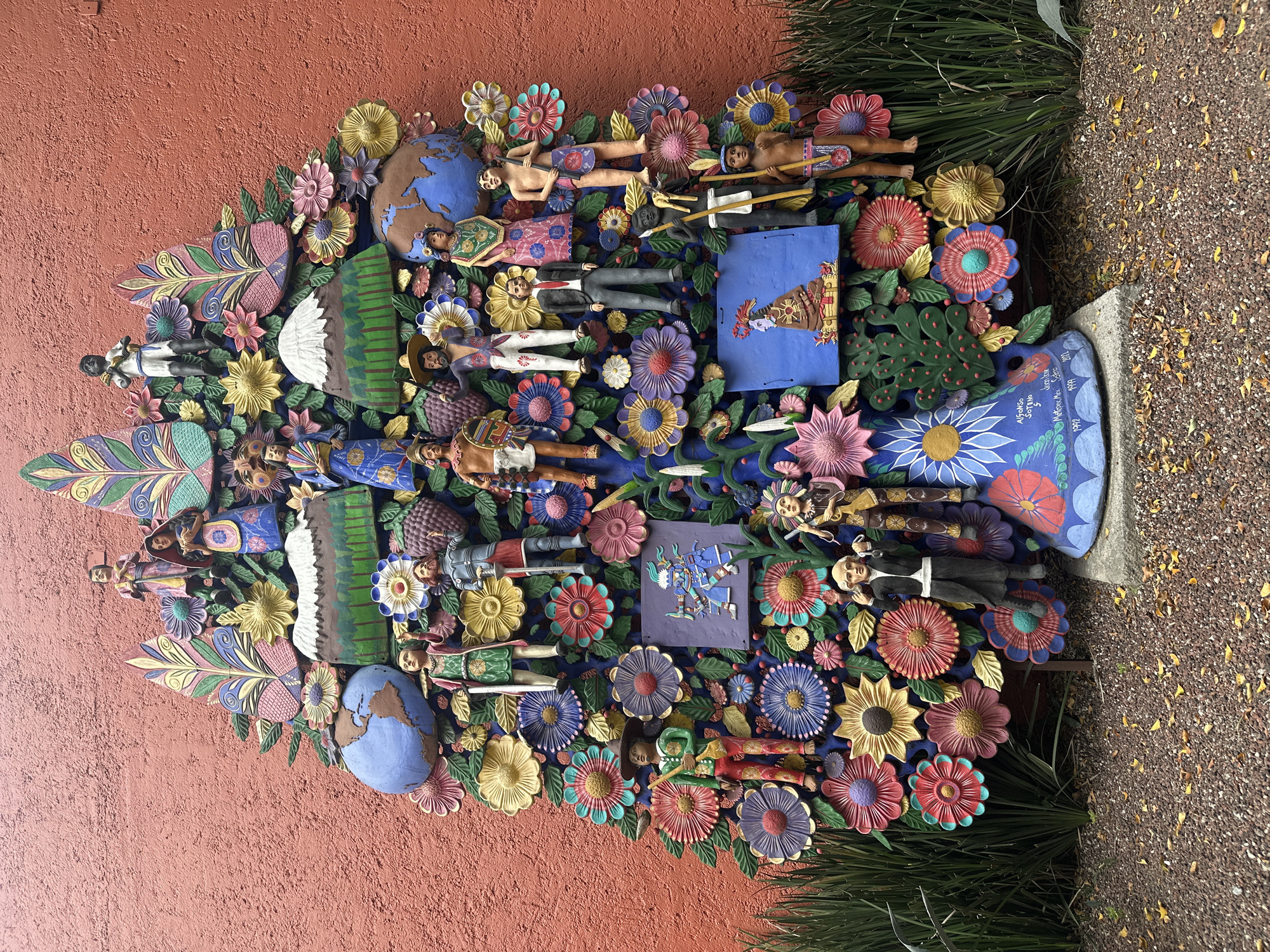 The image shows a large, colorful outdoor artwork on a brick wall in Mexico City, Mexico. The artwork is composed of numerous intricately carved and painted figures and flowers arranged in a tree-like shape. The figures depict people in historical and cultural attire, along with representations of Earth and Mexican flora. The piece is signed, “Alfonso Soteno Mex 1991, Jennifer Martinez Soteno 2012,”. The environment includes a brick wall that serves as the background and surrounding foliage and stone, suggesting the piece is in a public outdoor location.