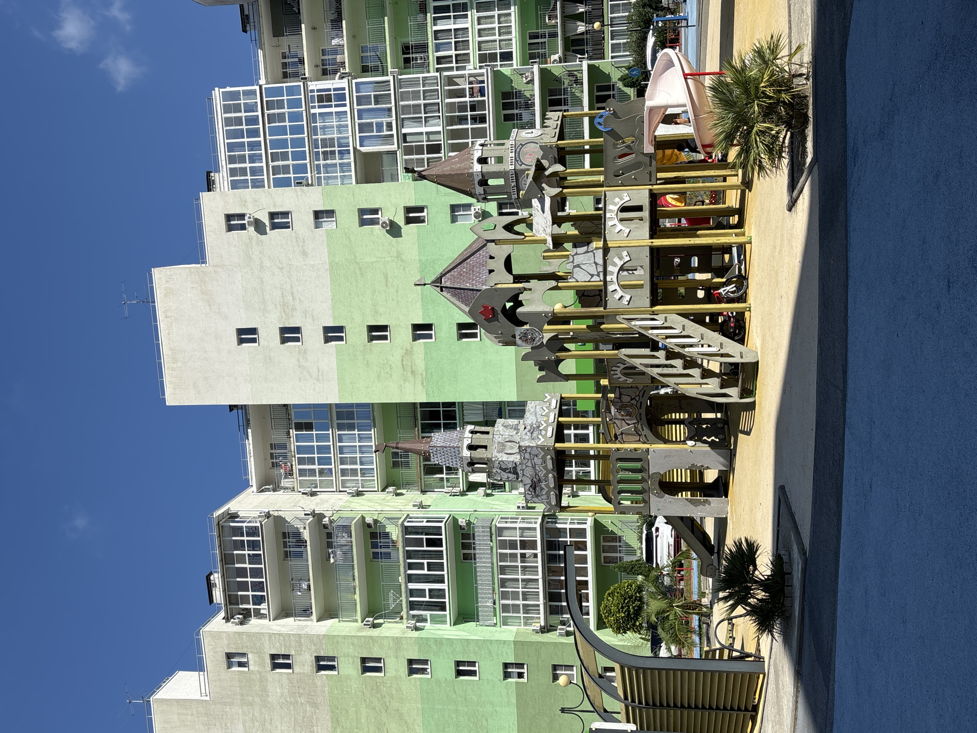 In Razdolnoye, Russia, a modern residential complex dominates the background with its light green and white-accented buildings featuring numerous balconies and windows. In the foreground, a colorful, castle-themed playground is set up on a sandy surface. The playground includes a slide, climbing structures, and decorative towers. Several palm trees are scattered around the playground area. The sky is clear and blue.