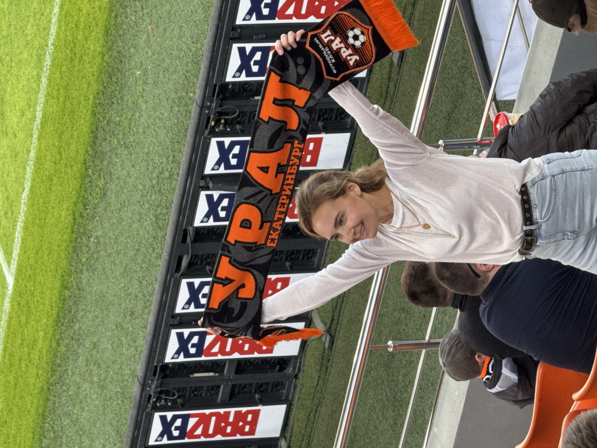 The image depicts a woman in the stands at what appears to be a soccer stadium in Yekaterinburg, Russia. She is holding up a black and orange scarf that displays the word "URAL" in large, orange Cyrillic letters. Below "URAL", the name "Yekaterinburg" is printed in smaller letters. The scarf also features a soccer ball emblem.
The woman is wearing a long-sleeved white shirt and light blue jeans with a dark belt. She is smiling and looking towards the scarf. Other people are visible behind and beside her in the stands. The background includes part of the soccer field and a digital advertising display with the word "BROZEX" repeated. The railings of the stadium seating are also visible.