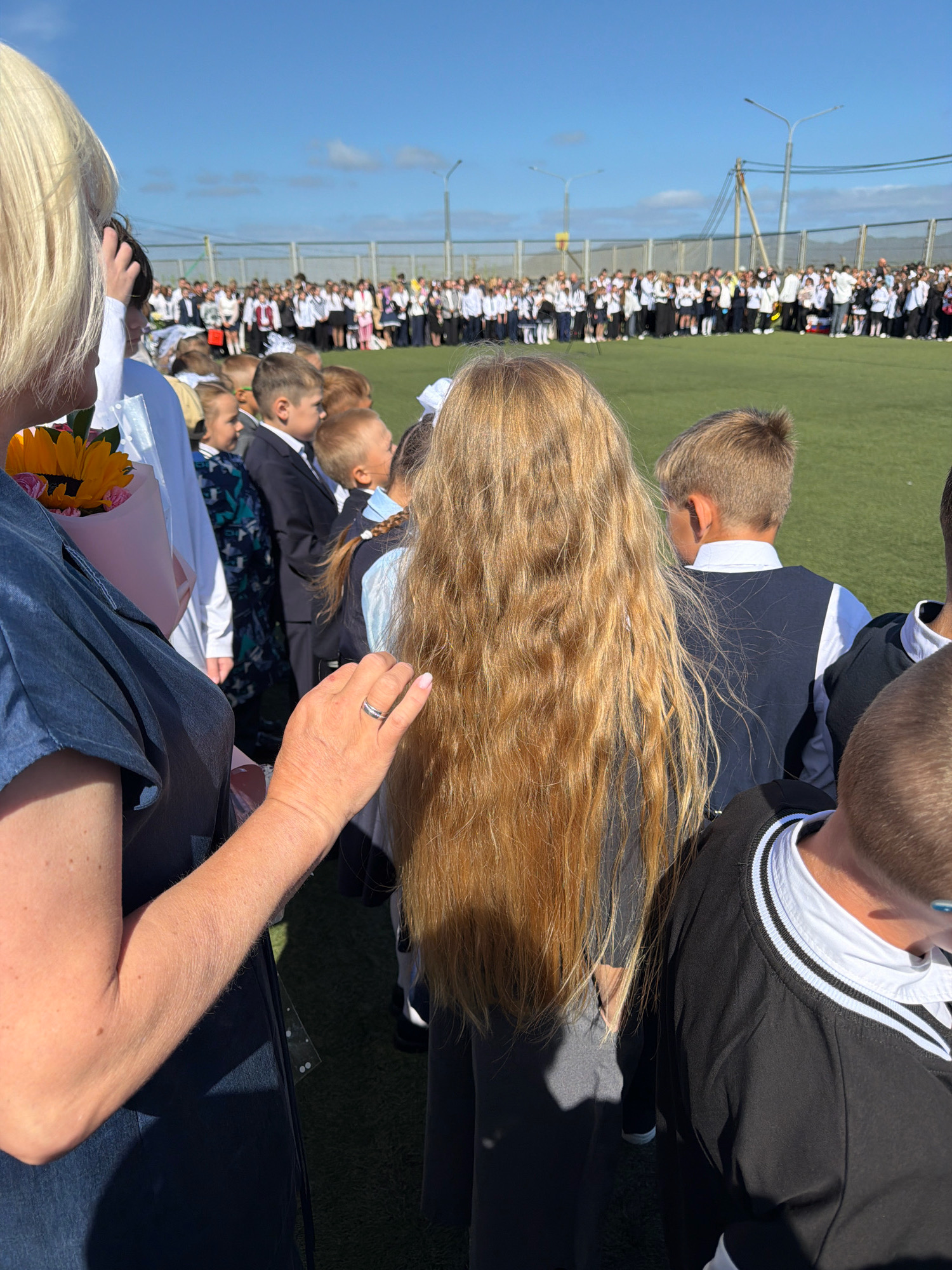 In Yuzhno-Kurilsk, Russia, on a clear day under a bright blue sky, a large gathering of schoolchildren in uniform stands in rows on a green field, likely a sports field. The children are predominantly wearing dark blazers or vests over white shirts, and some are holding flowers. The foreground of the image shows the backs of several people, including a woman with blonde hair, her arm visible wearing a ring, and a young girl with long blonde hair. A bouquet of yellow and pink flowers is held by someone in the foreground. The background reveals a fence and utility poles, suggesting an outdoor public space within the city. The overall scene depicts an event, possibly a school assembly or holiday celebration, characterized by orderly rows of uniformed students.