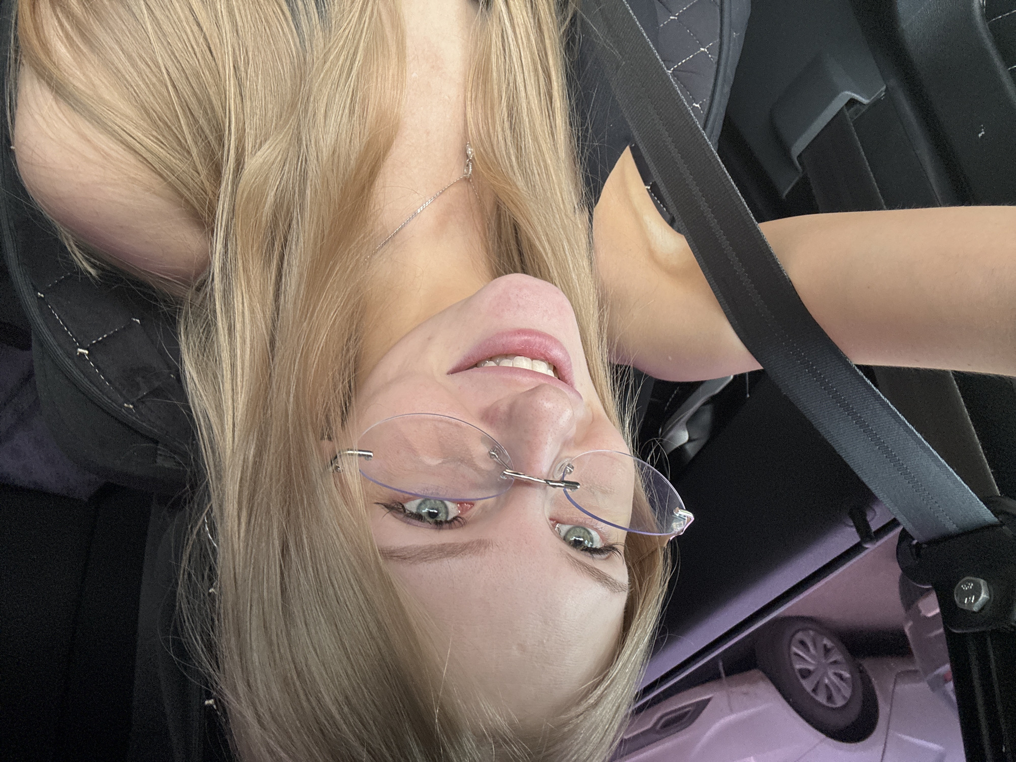 A close-up, upside-down selfie of a young woman with blonde hair and green eyes, wearing glasses, is captured inside a car. Her hair is long and light brown, falling around her face and shoulders. She is wearing a black top and a seatbelt, suggesting she is traveling. The setting appears to be daytime, with the interior of the car visible, including a seatbelt mechanism and a glimpse of a car wheel through the window. The woman's expression is a slight smile, and the overall mood is casual and possibly playful, given the unusual selfie angle. No visible text is present in the image.